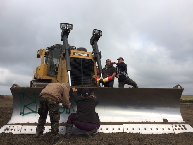 An indigenous activist locked to an excavator takes a drink of soda, while two others spray paint "Black Snake Kills" on the blade. "Black Snake" refers to an old Lakota prophecy, about a black snake that would come to the prairie with the ability to destroy the world — or unify it.