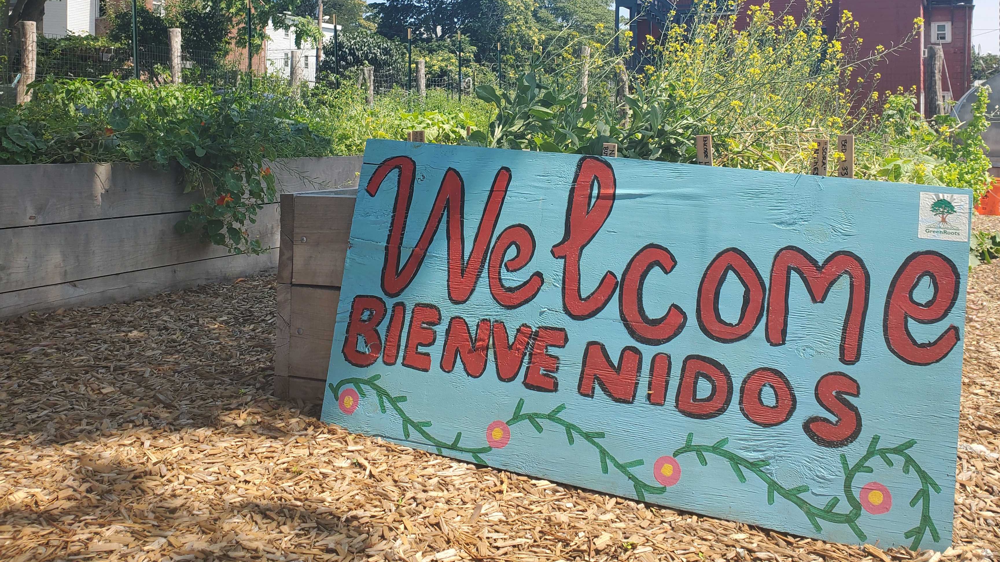 A sign resting on a wooden garden bed overflowing with green plants reads "Welcome, Bienvenidos."