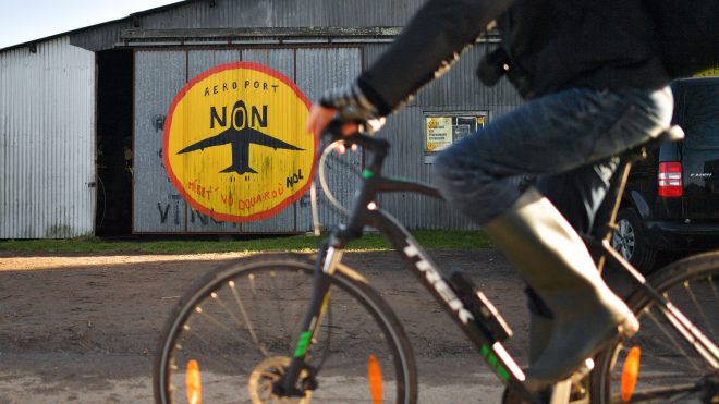 Photo of a person biking past a barn painted with an airplane, part of a utopian community protesting an airport in Western France.