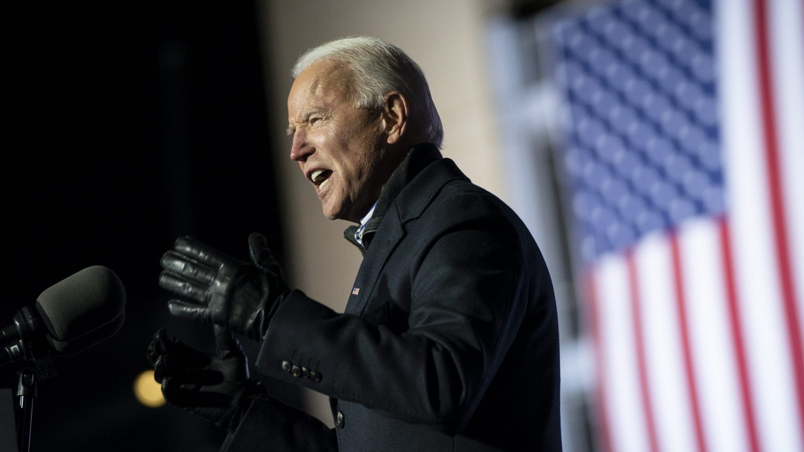 A photo of Joe Biden at a podium speaking. He is wearing a dark coat and dark gloves.