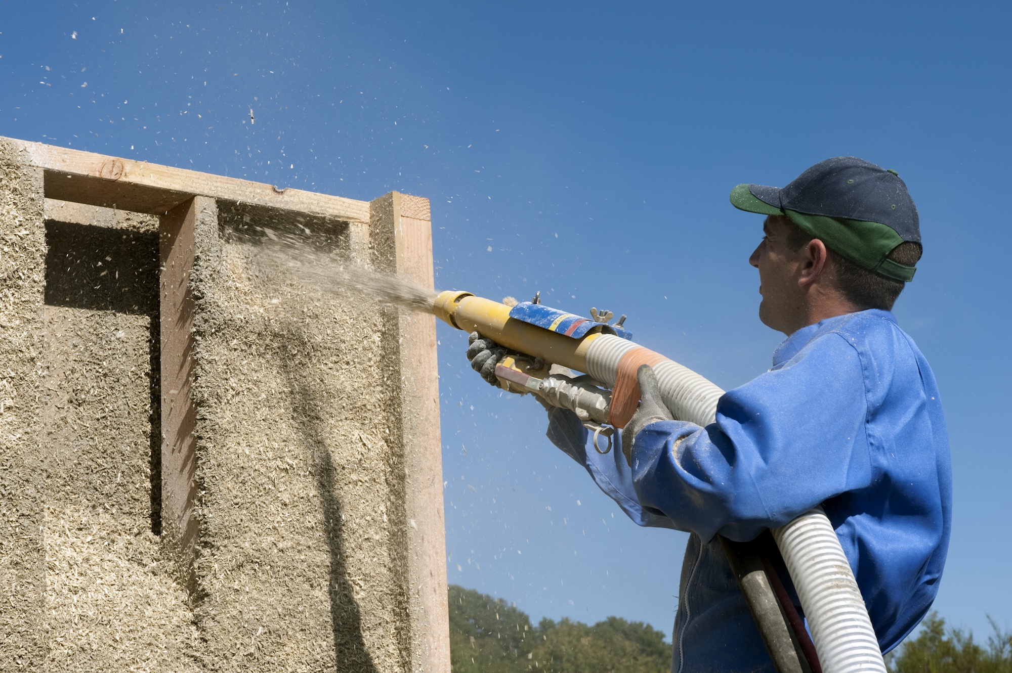 A man in a blue shirt and baseball cap holds a spray hose blasting out a mixture of small bits of hemp into a wooden building frame