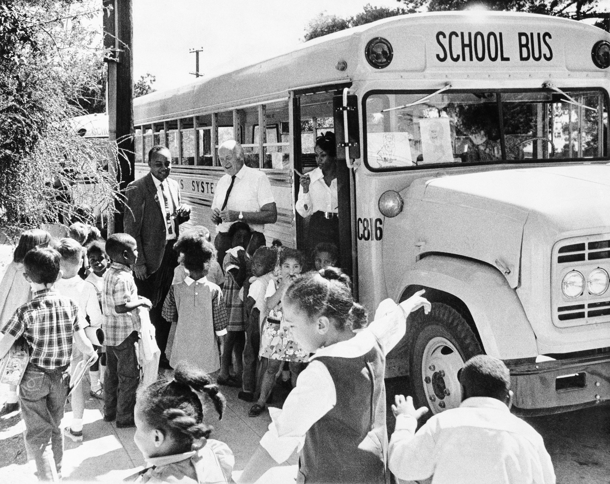 A black and white photo of Black children heading for a school bus while adults supervise