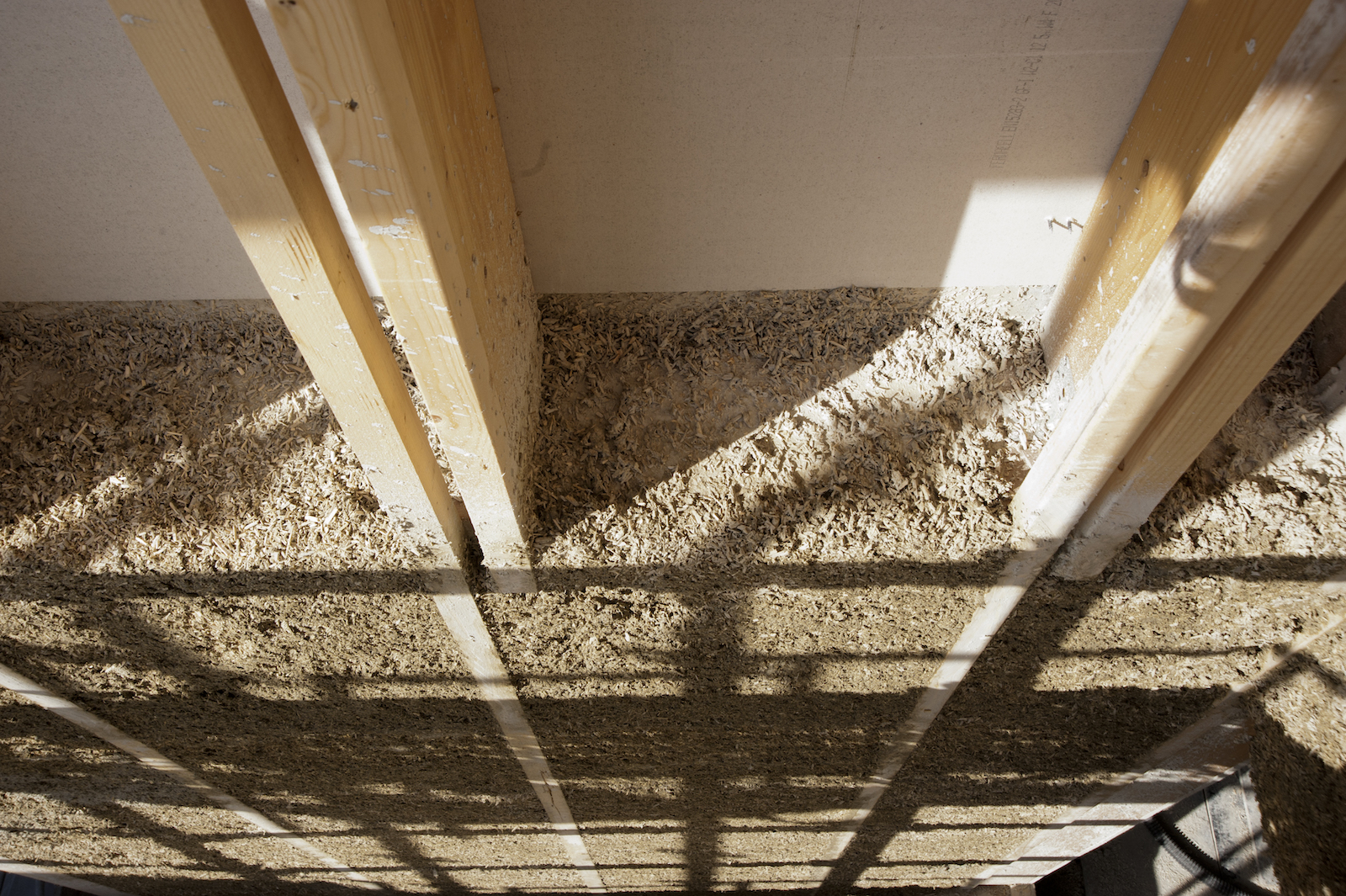 a photo of a top-down view of the inside of a wall. Long wooden beams run in parallel. In between, grayish rectangles of "hempcrete" fill in the gaps. It is porous-looking like a sponge.