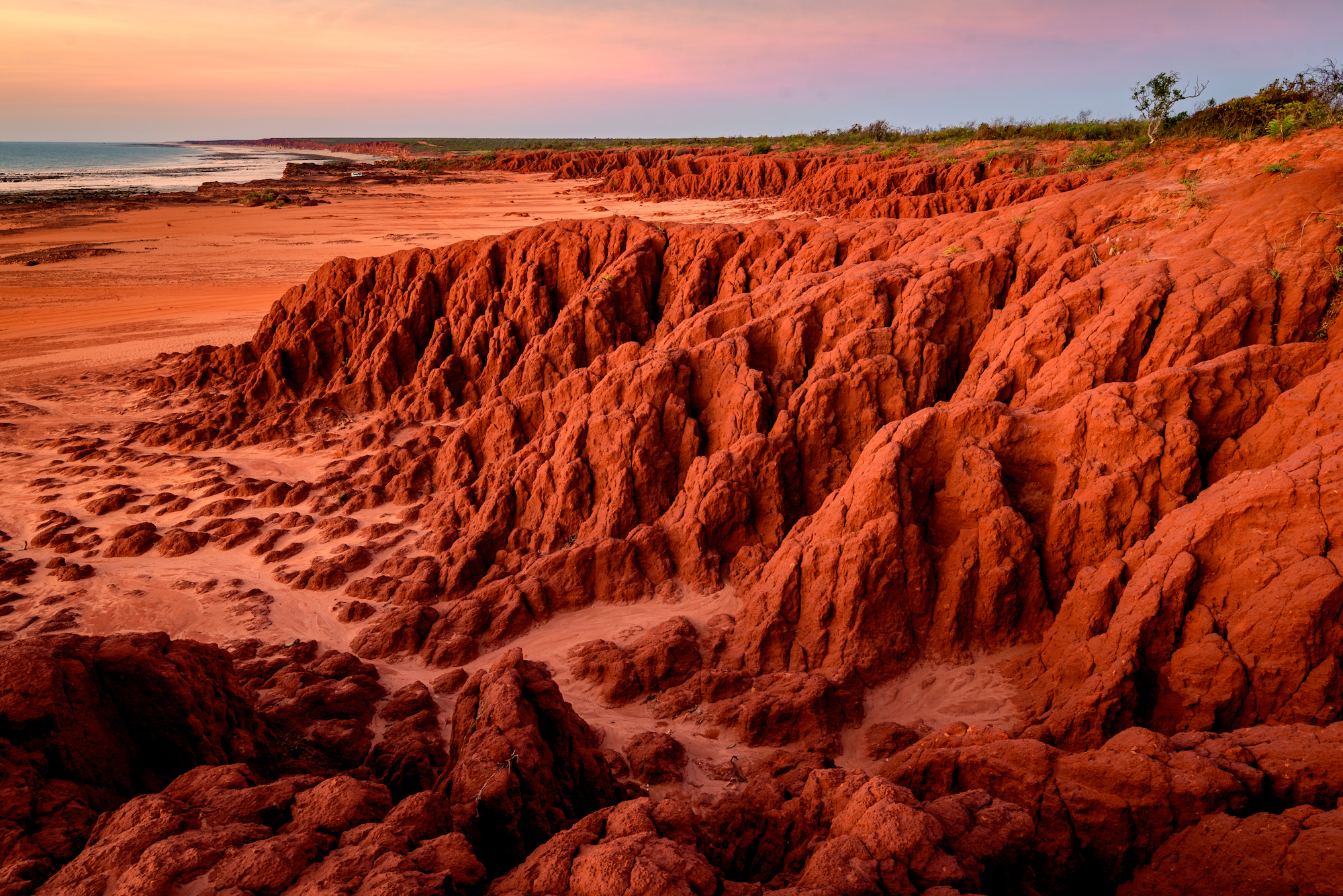 deep red, rocky cliffs stretch from the right side of the screen, melting into sandy beach and blue water