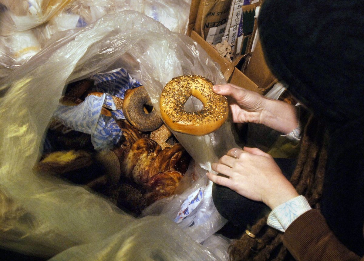 A person holds a seeded bagel above a garbage bag filled with baked goods.