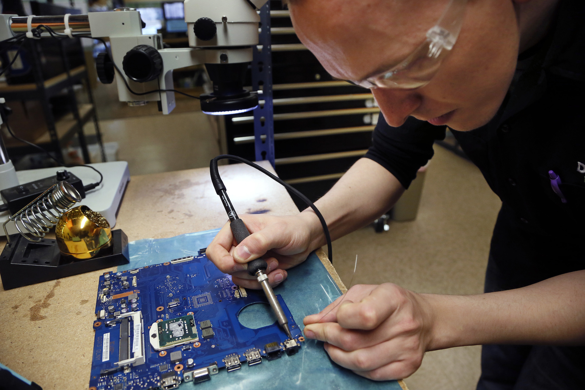 A photo of a man wearing goggles leaning over blue laptop motherboard while holding a tool