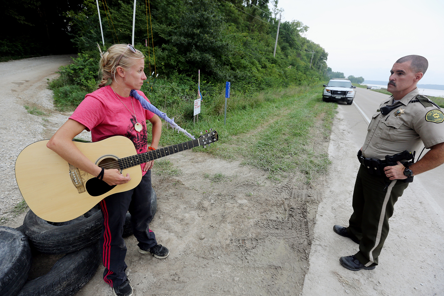 Jessica Reznicek, holding an acoustic guitar, stares at a uniformed officer while standing near a tree-lined road