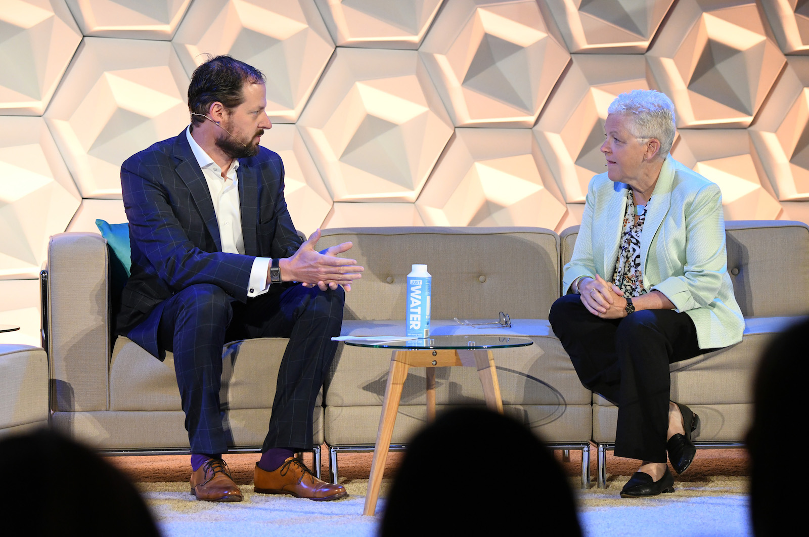 a man in a blue suit sits on a couch in conversation with a woman with short white hair. They are speaking on stage and the back of the audience's head is just visible at the bottom of the image.