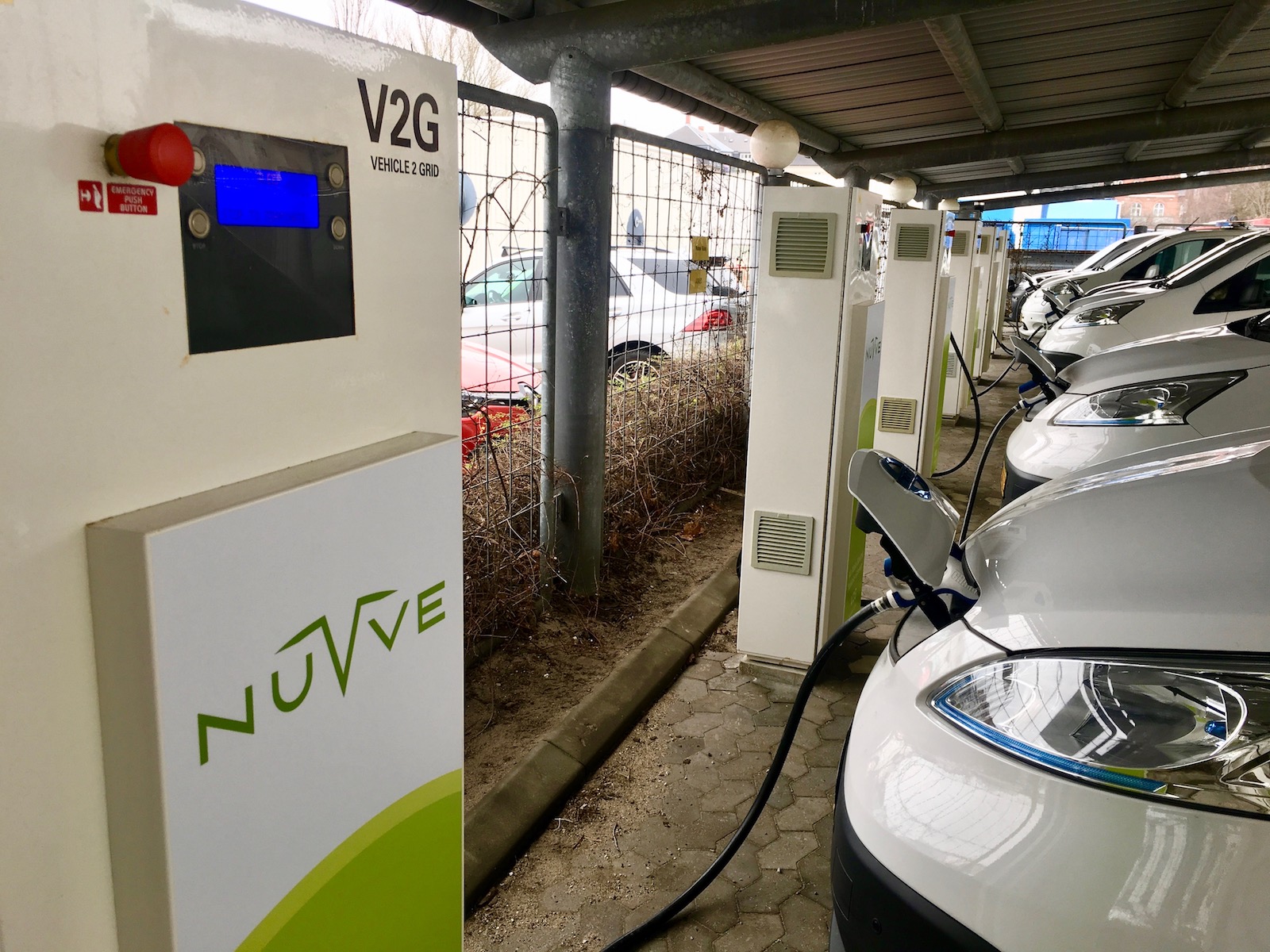 A row of shiny white cars are plugged into green and white wall chargers labeled "Nuvve V2G"