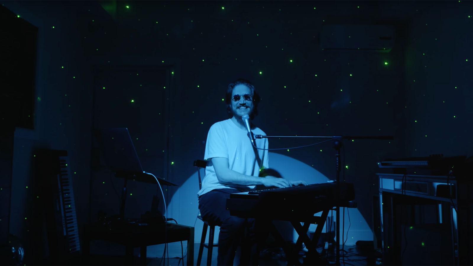 A man wearing sunglasses plays the keyboard while a blue spotlight shines on him, with a starry sky projected in the background