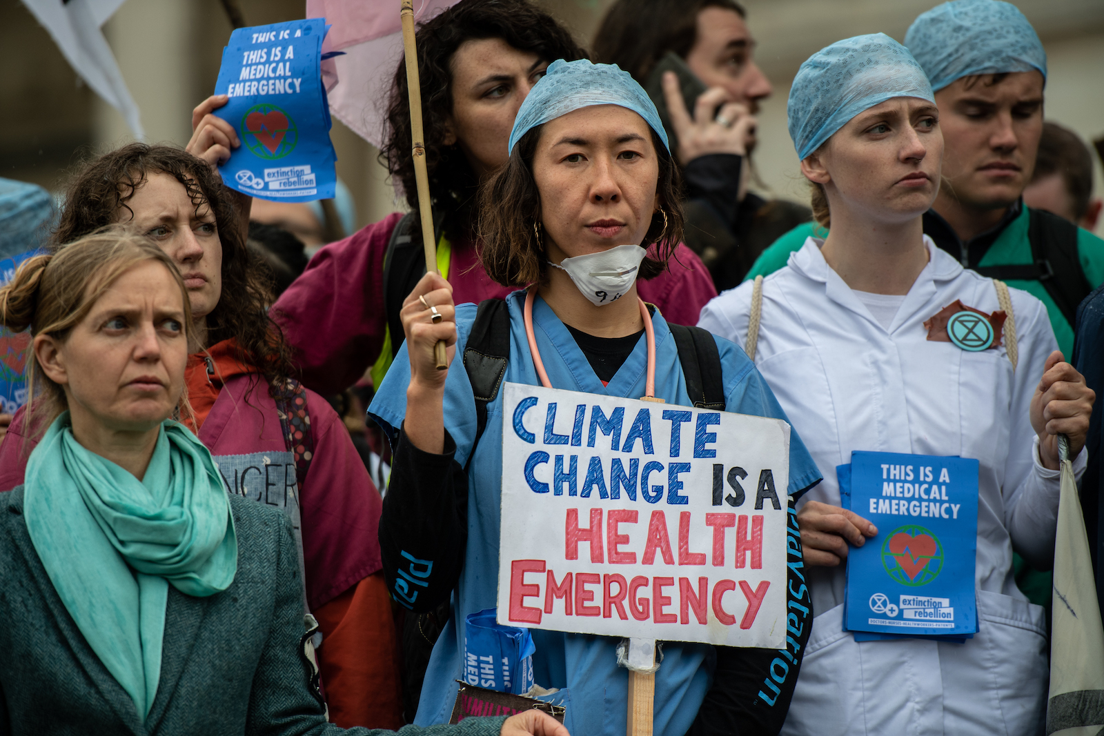 a group of protesters, with a woman in a scrubs and a surgical mask standing in the center. She holds a sign that reads, "climate change is a health emergency"