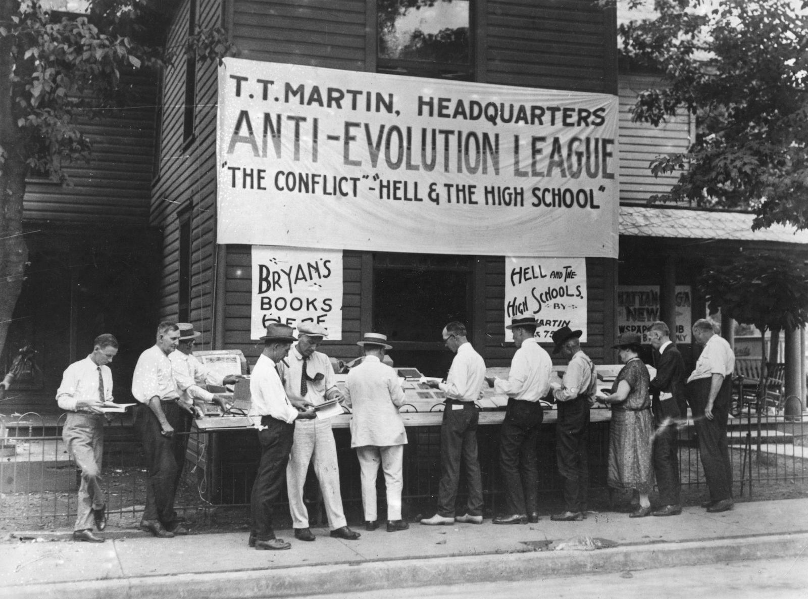 a large sign for the "anti-evolution league" hangs above a black and white scene of people in front of a book stand