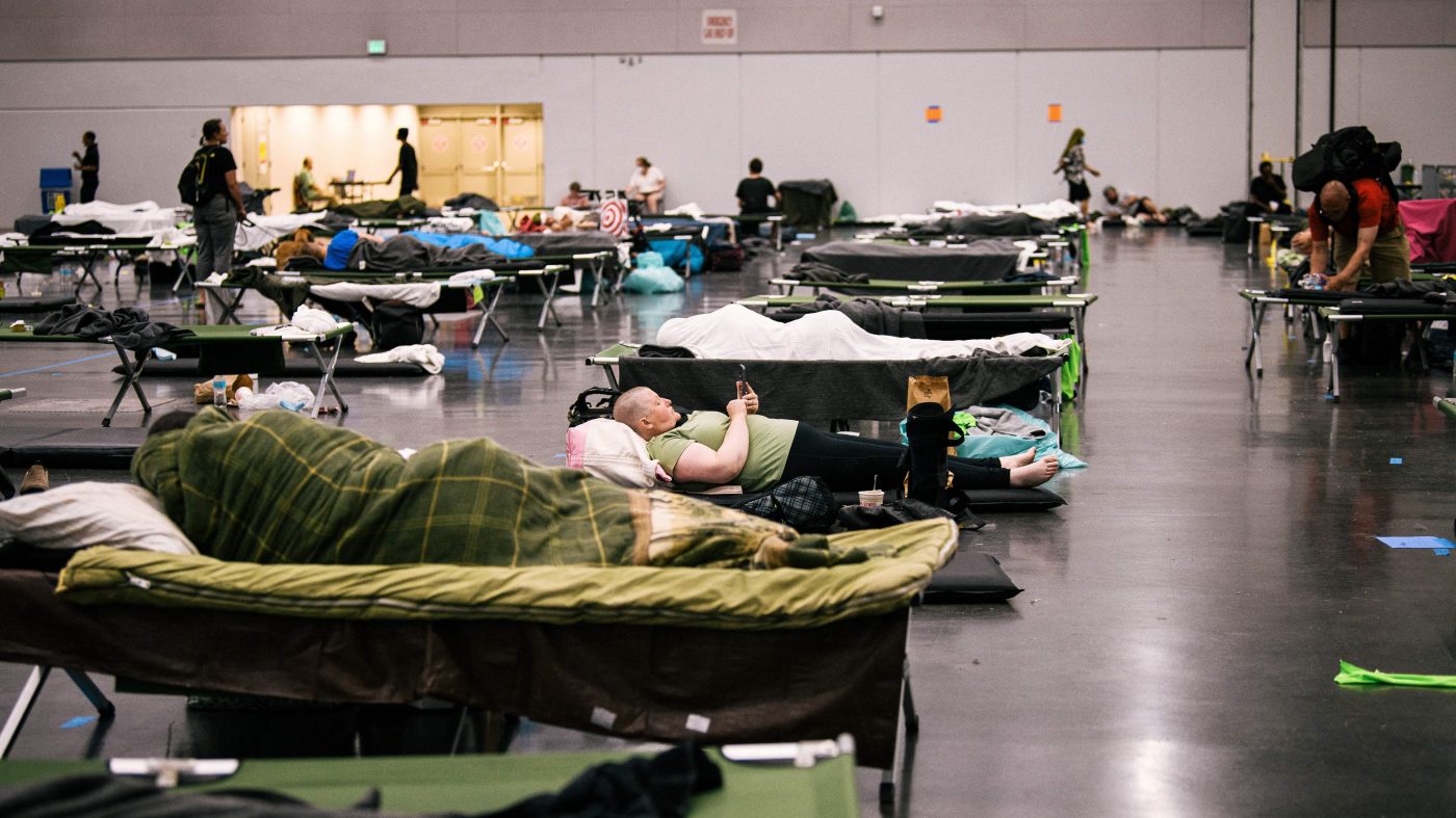 People rest on cots with blankets in a large room.