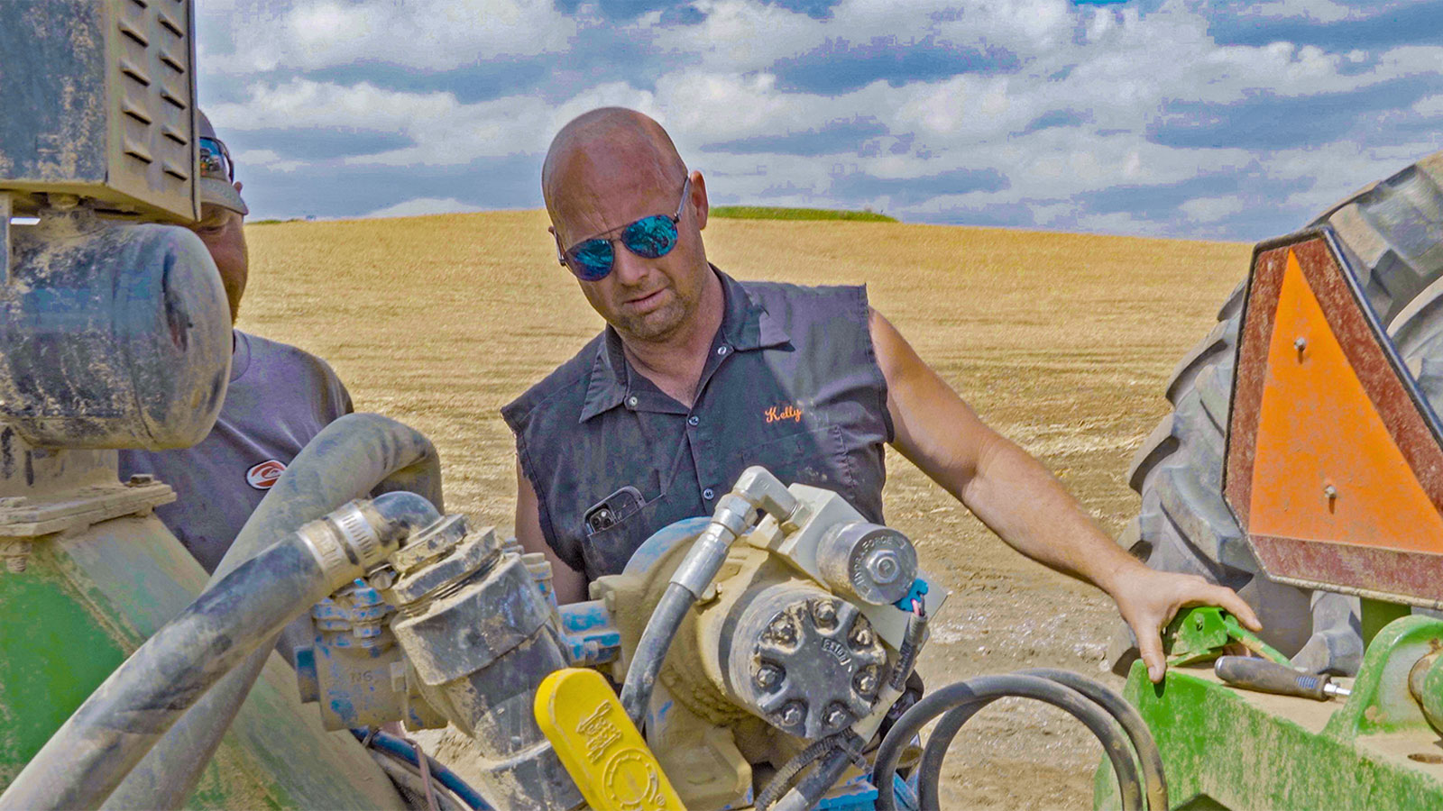 A bald man in sunglasses and a cut-off shirt looks over a piece of farm equipment