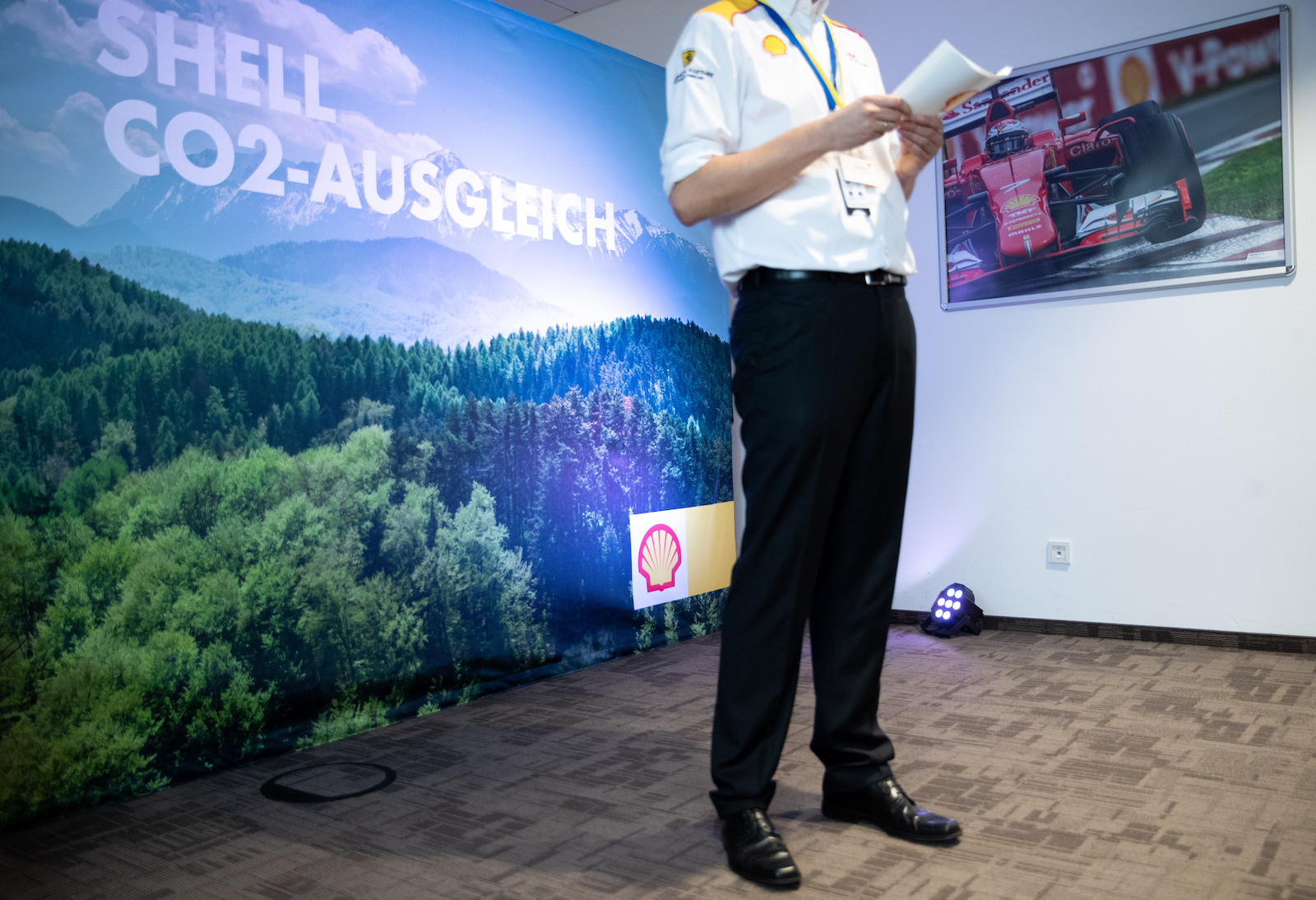 a man in a white shirt and black pans stands in front of a large mural covered in trees promoting a tree-related CO2 offset by Shell company