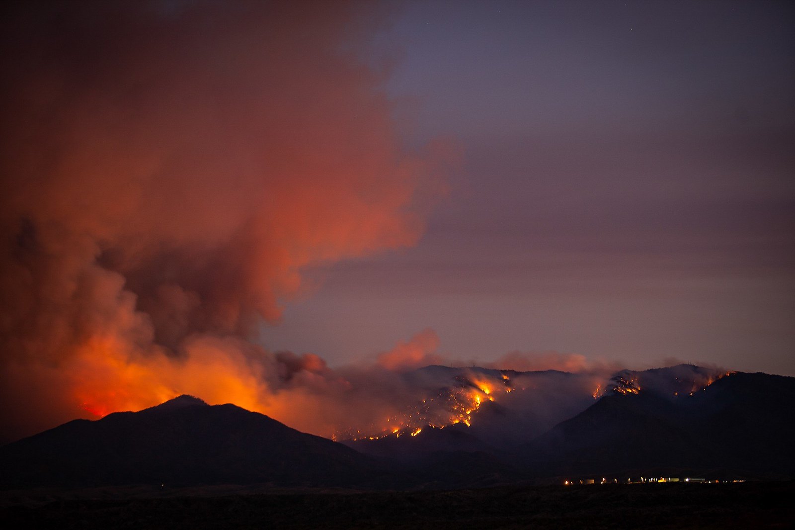 a wildfire with smoke peeks over a darkened hill against a sunset landscape