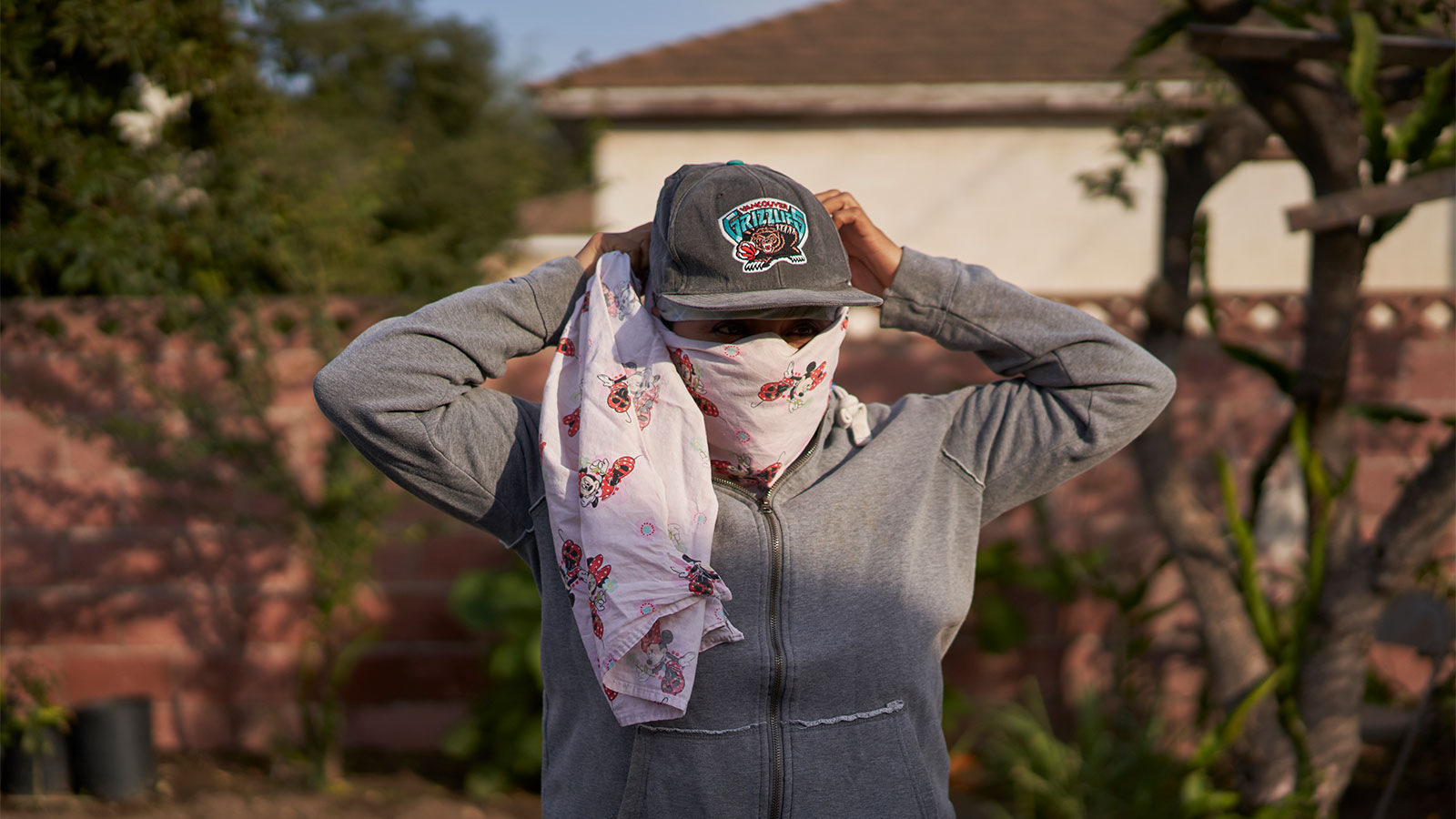a woman ties a bandana around her face while wearing a hat and standing in front of a sunny house