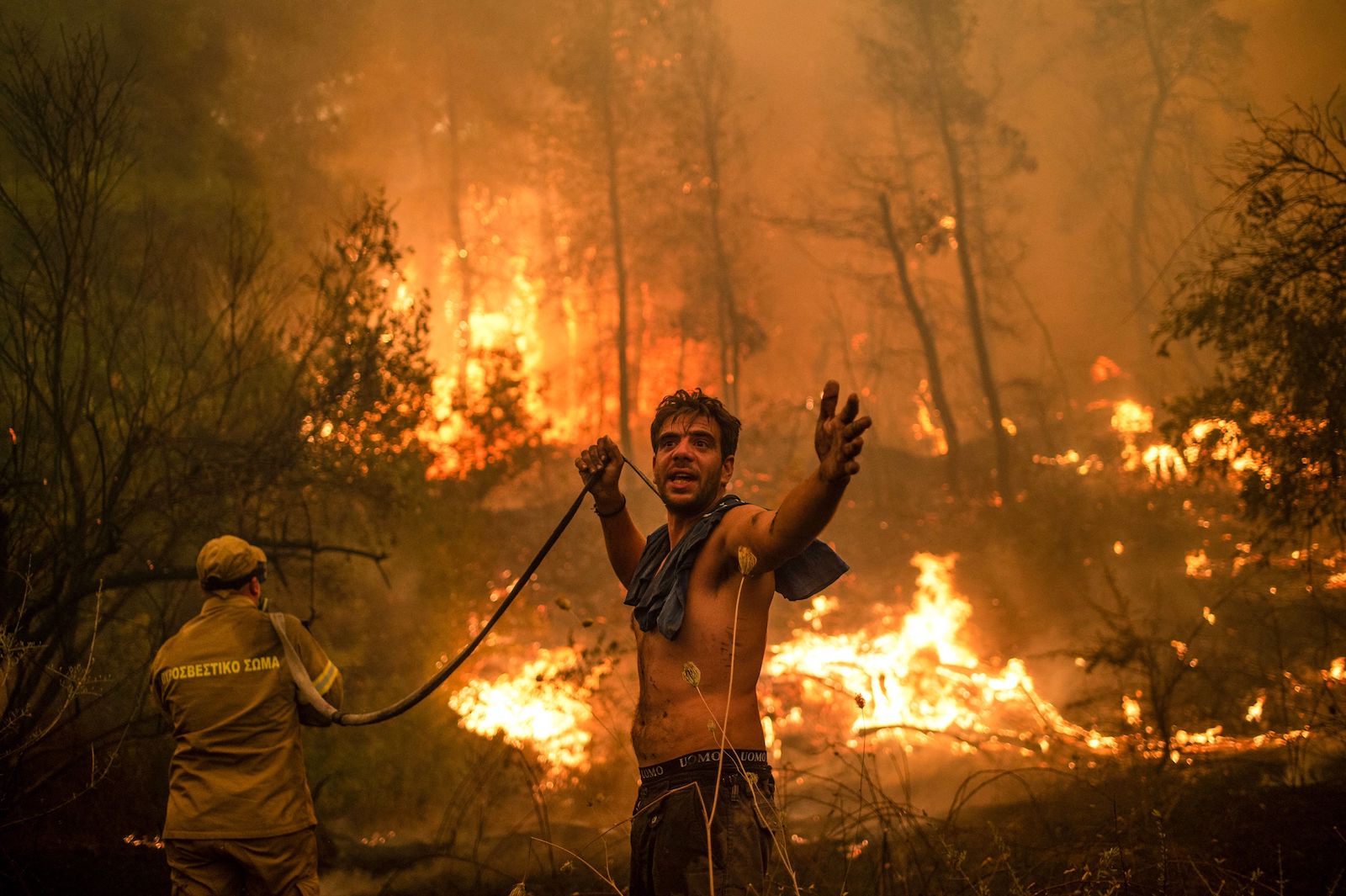 a shirtless man holding a fire hose gestures at the viewer as wildfires blaze behind him