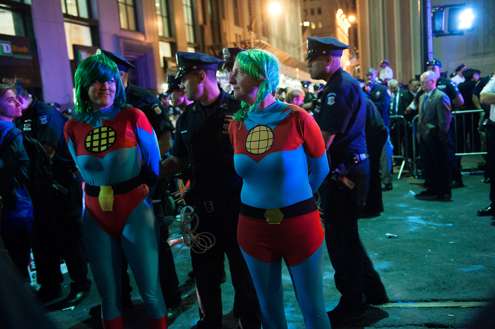 a night scene: two women in blue tights, green wigs, and red shorts are handcuffed by police on the street