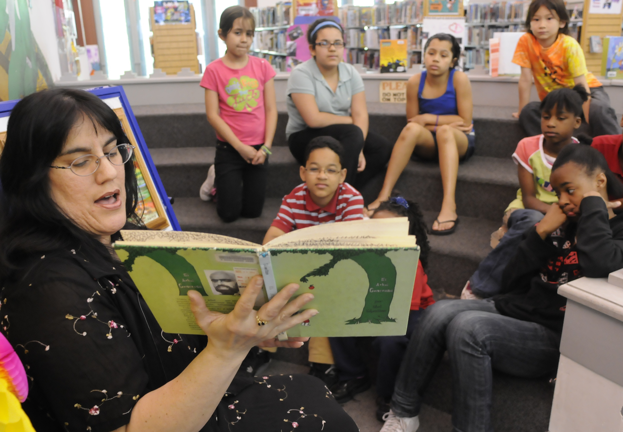 a woman with glasses holds open a copy of the giving tree with a green cover. Kids sit on risers in the background looking at the book