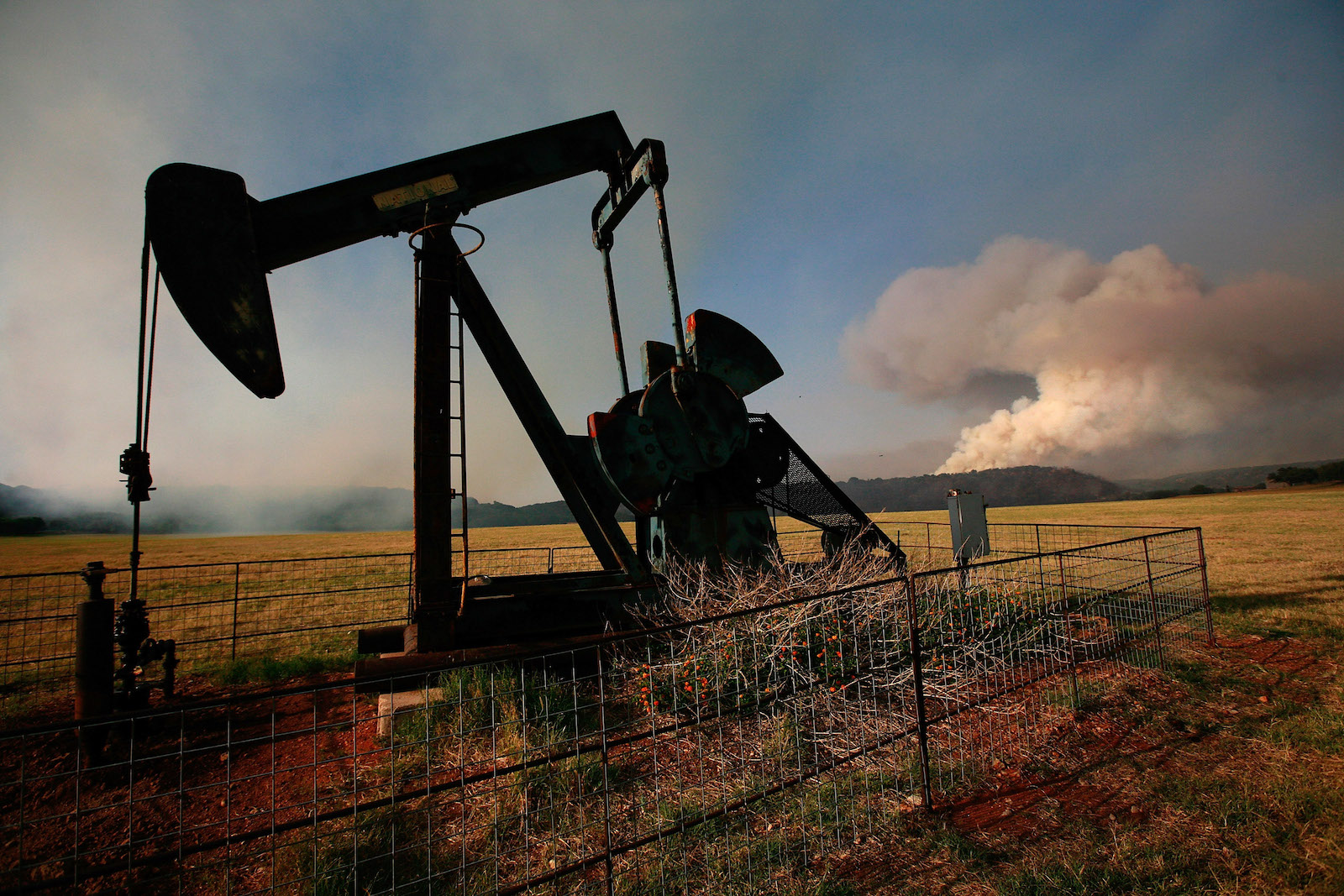 an oilfield pumpjack is silhouetted against a dry landscape. In the distance, a large white plume of smoke billows into a blue sky