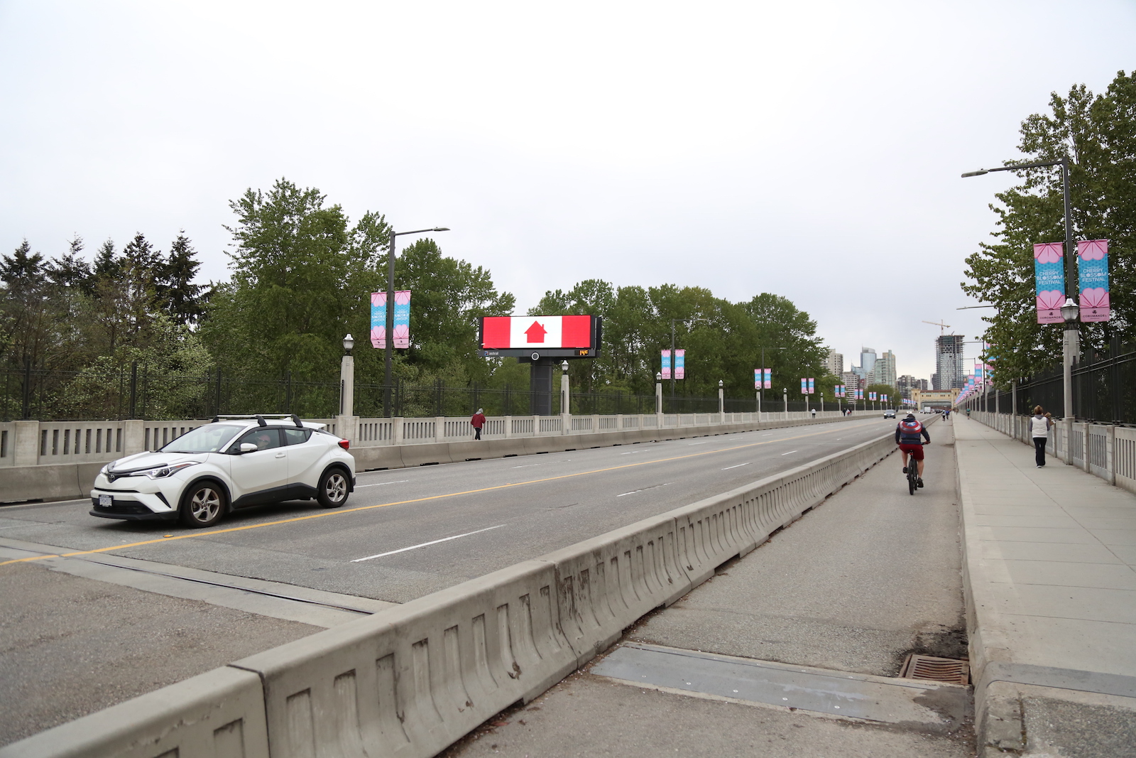 a single white car zooms on the other side of a protected bike lane with one rider. A billboard with an adaptation of the Canadian flag glows on the side of the road