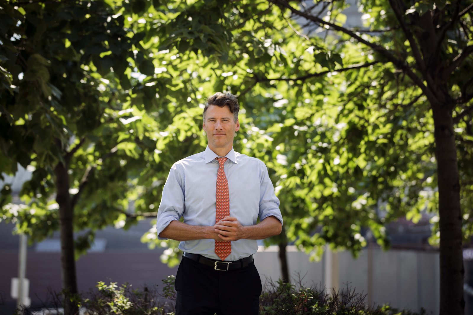 a man in a button-up shirt and a coral tie stands in a tree-filled area