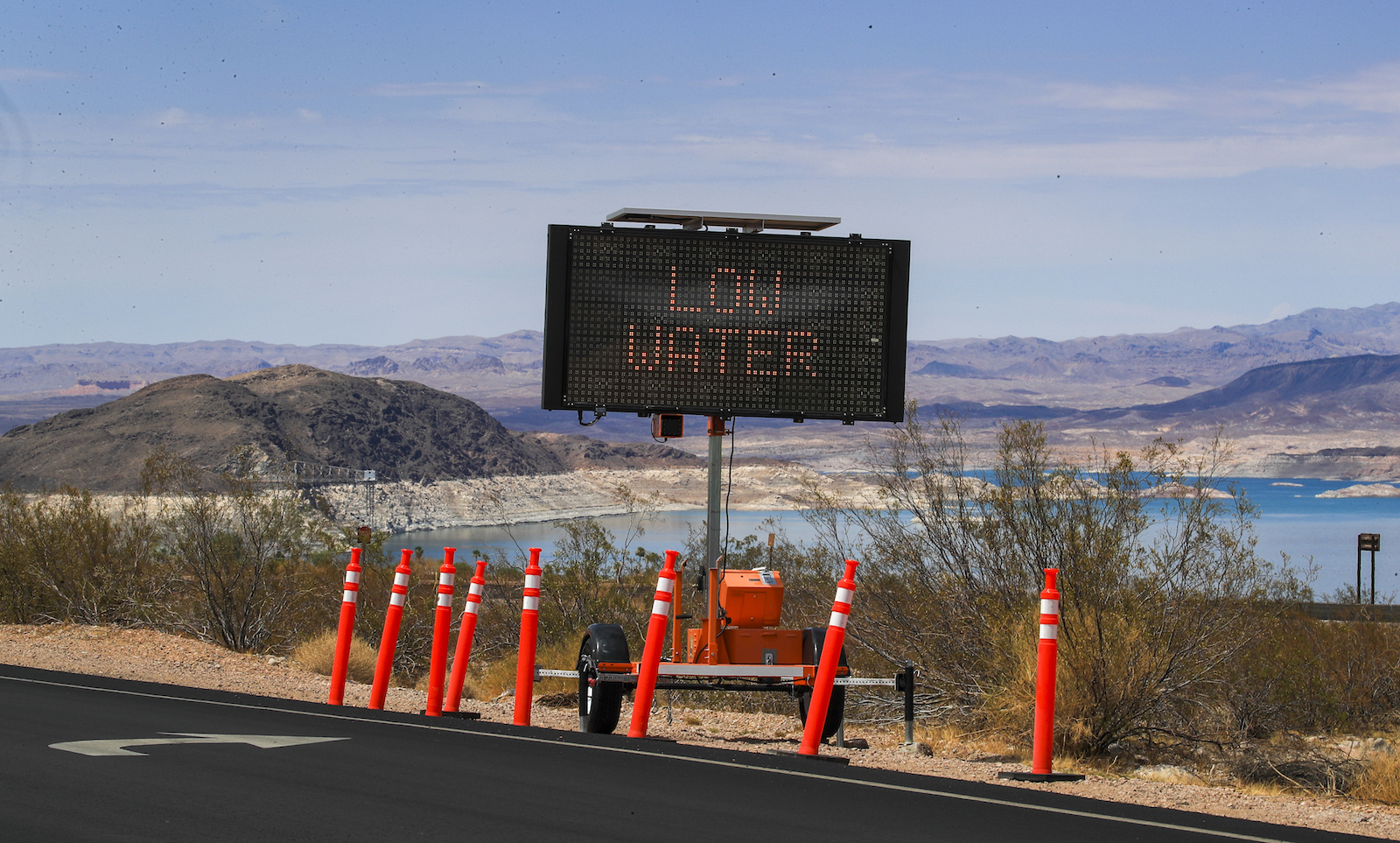 a large light-up sign near the side of a highway says low levels. The sign is surrounded by orange cones and water is visible in the background
