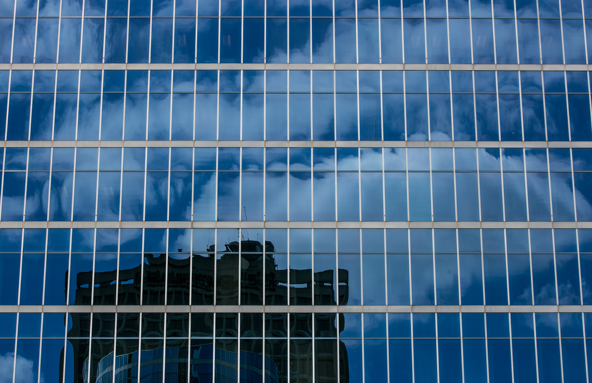 a blue glass building facade reflecting clouds, sky, and a nearby building