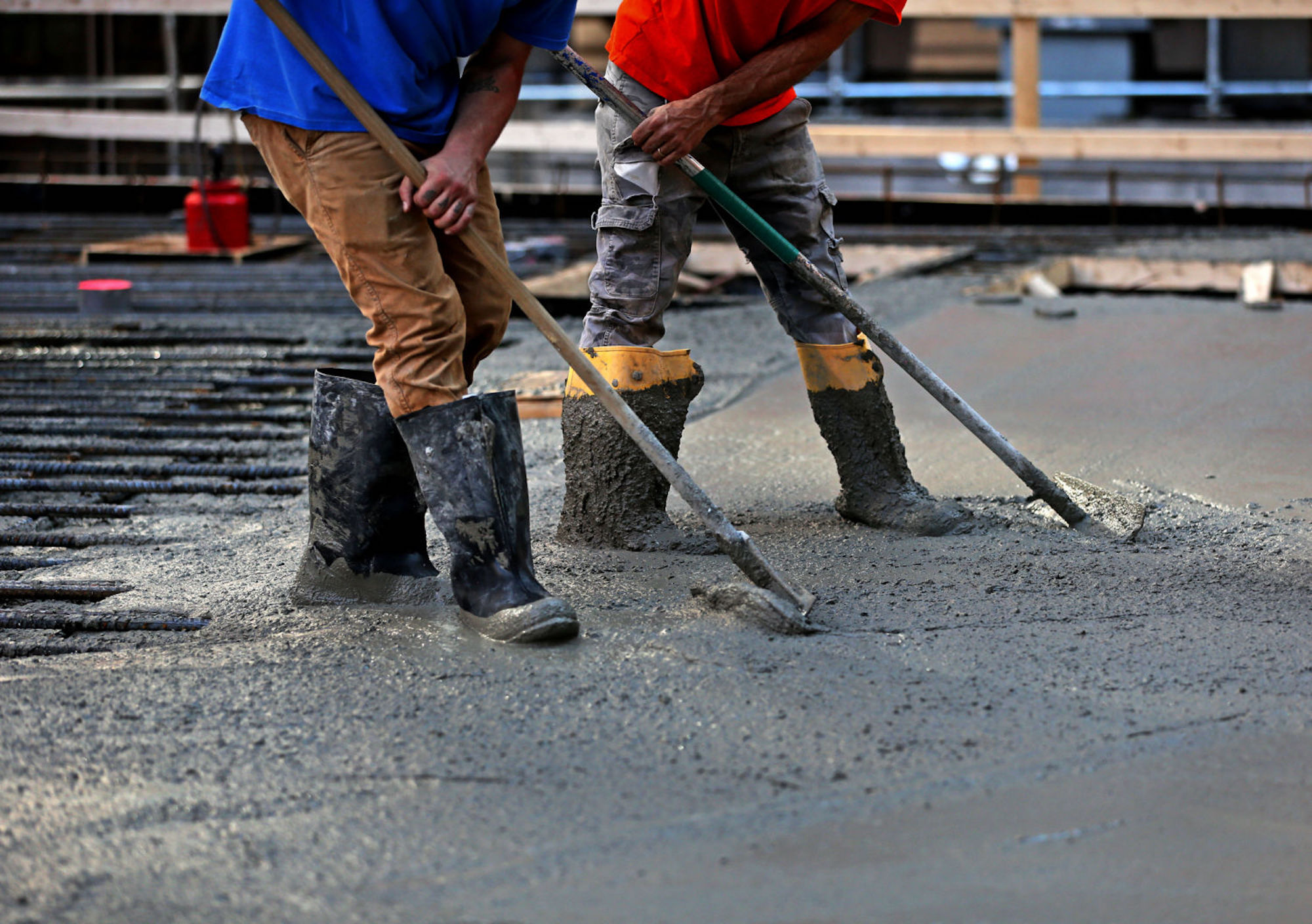 Two men, one wearing a red shirt with grey pants, and the other one a blue shirt and brown plants, use shovels to distribute fresh concrete over a street. They're also wearing construction boots.