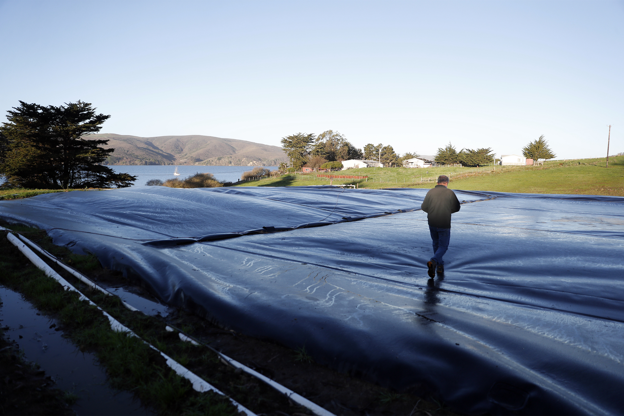 A man dressed with black plants and a black jacket walks over a giant black plastic bag, where methane is being stored. You can see mountains in the background, a clear, blue sky, and a couple of trees.