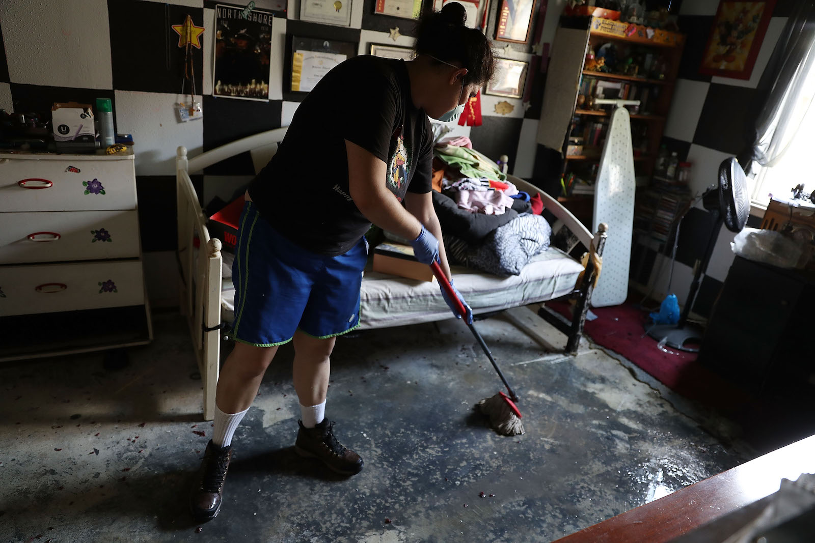 A woman with dark hair, wearing blue short and a black t-shirt, mops up floodwater in her bedroom in Houston, Texas following Hurricane Harvey in September 2017.
