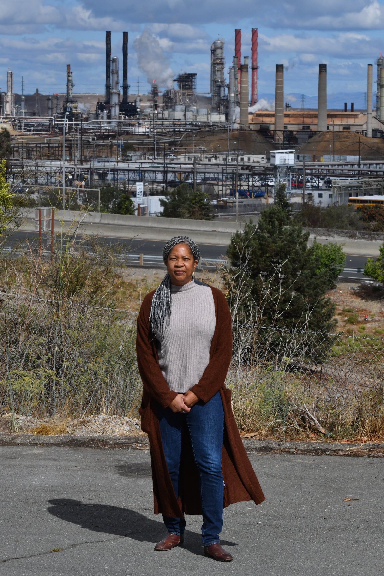 a woman in a gray shirt and red long coat stands in front of a refinery