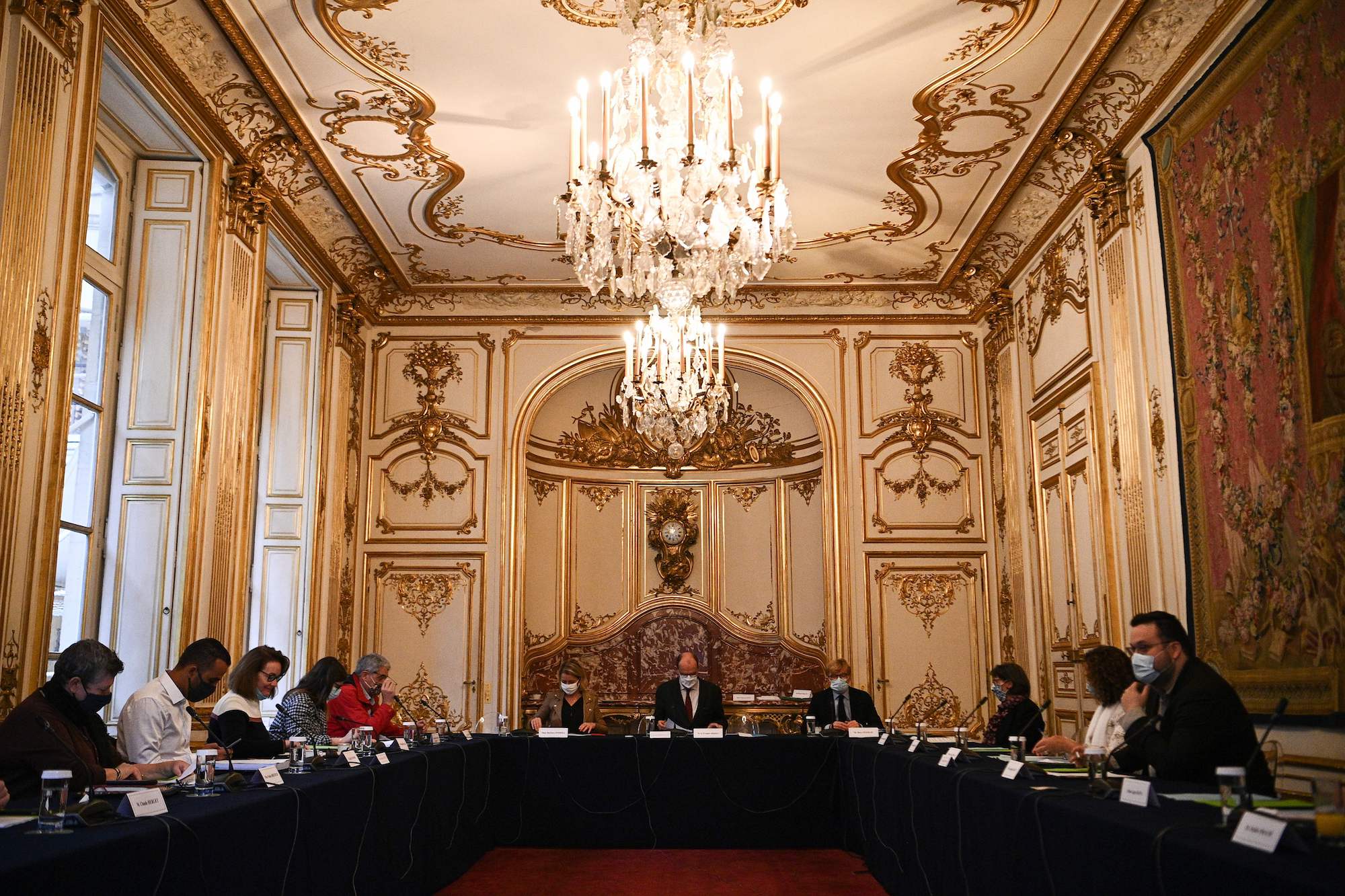 a gilded room with giant chandelier and long tables with people sitting at them discussing