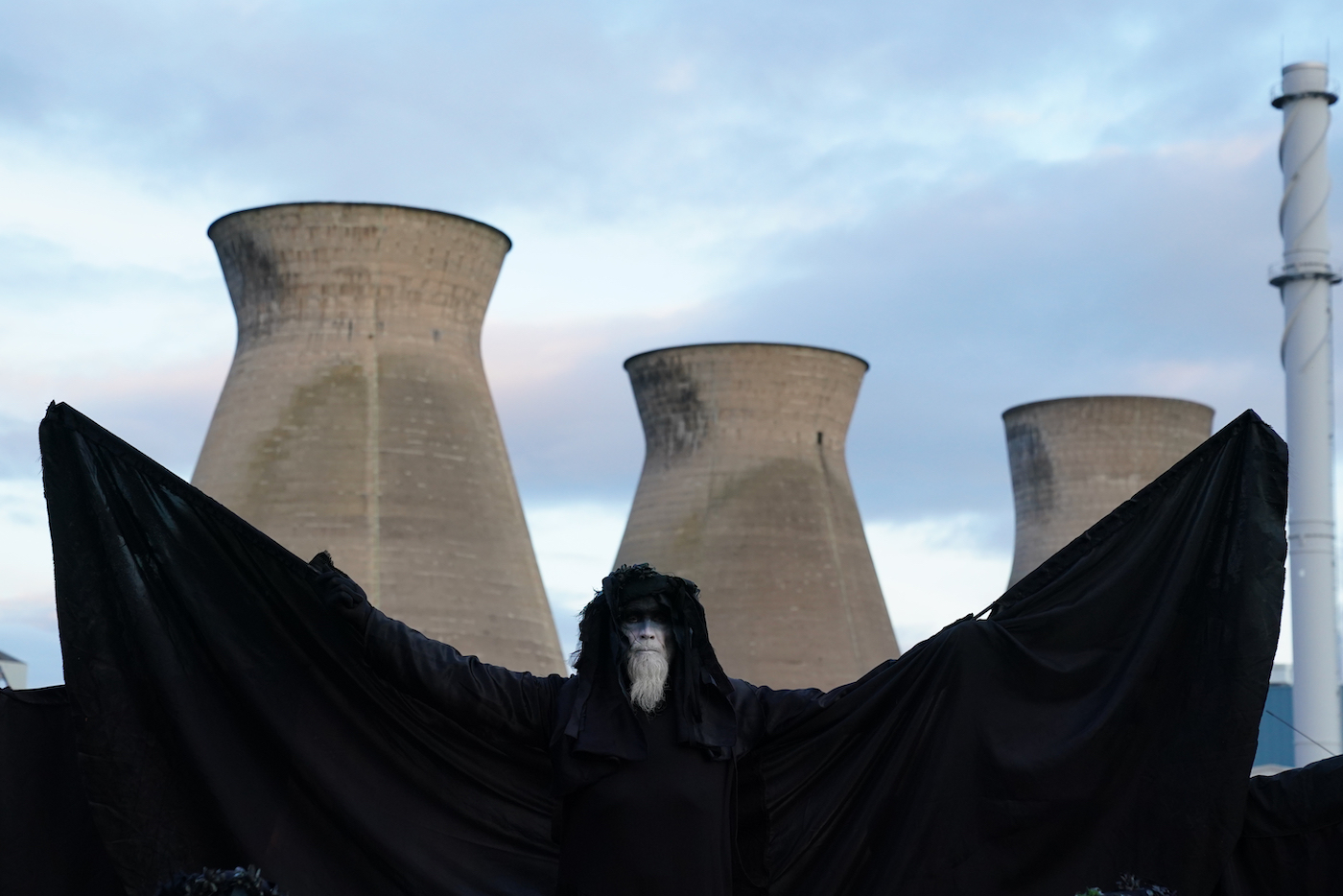 An activist in a black cape with arms outstretched like a bat stands in front of an oil refinery