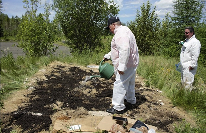Two men in white hazmat suits water a bare plot of soil.