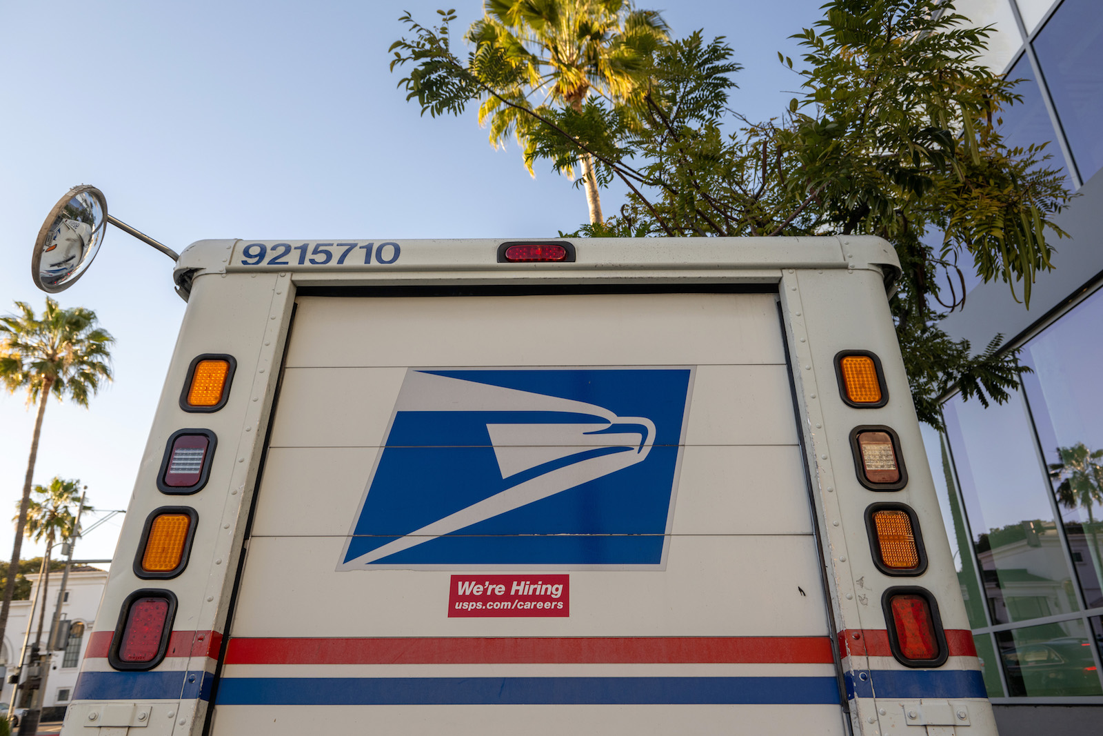 the back of a USPS mail truck with blue eagle logo and brake lights. In the background, palm trees and blue skies