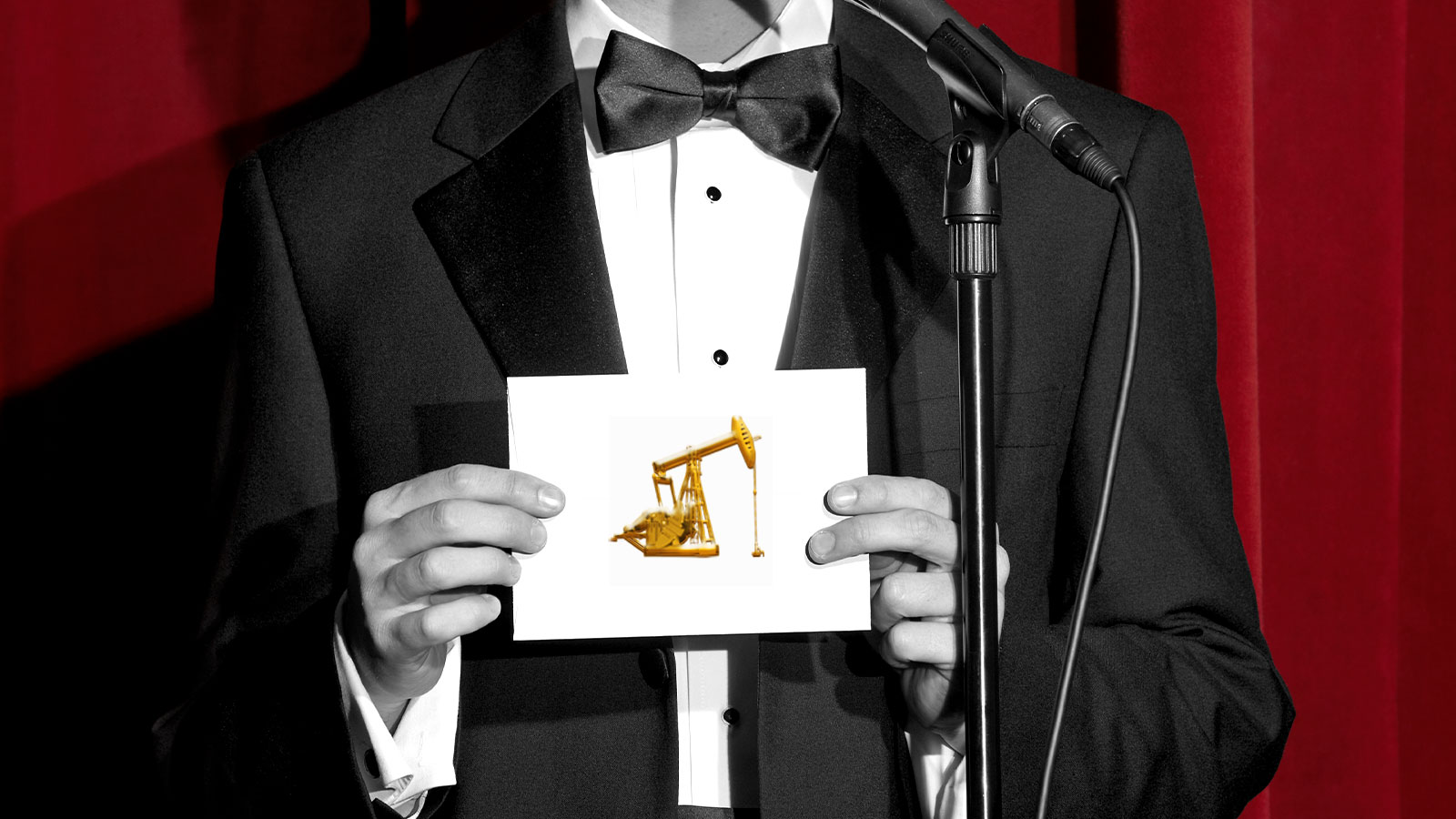 black and white photo of man in tux holding an envelope with a gold oil pump on it; red curtains in background