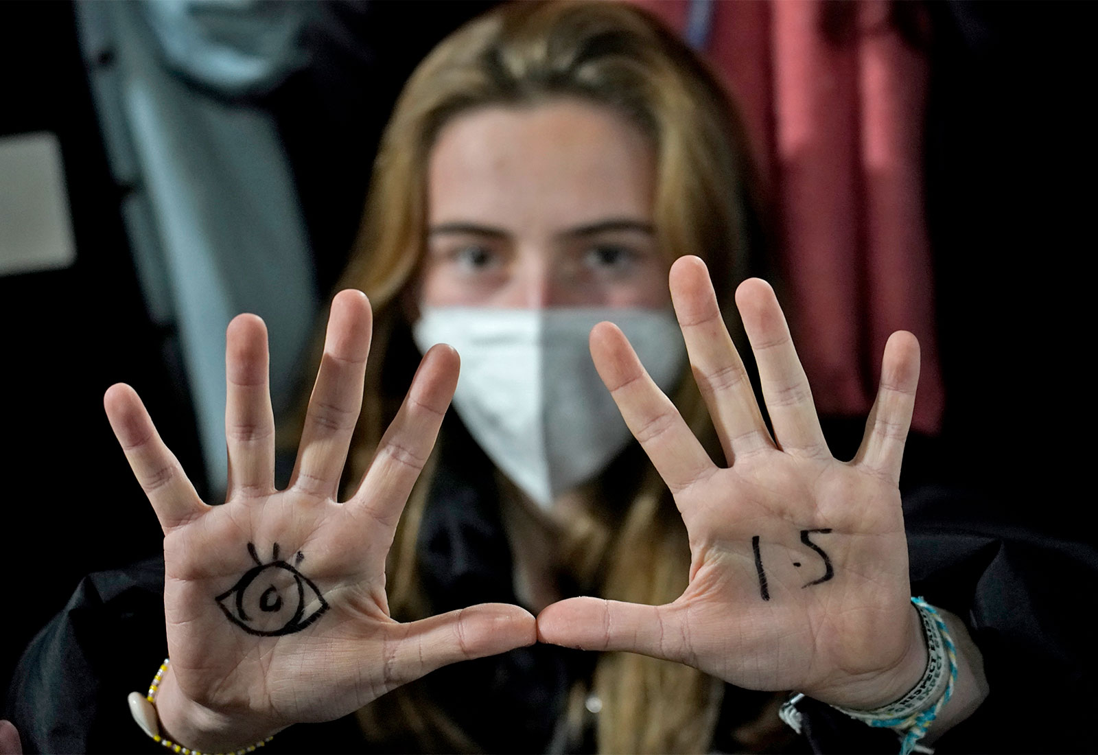woman wearing mask looking at camera holding up her hands to show an eye drawn on her left palm and 1.5 written on her right palm in black marker