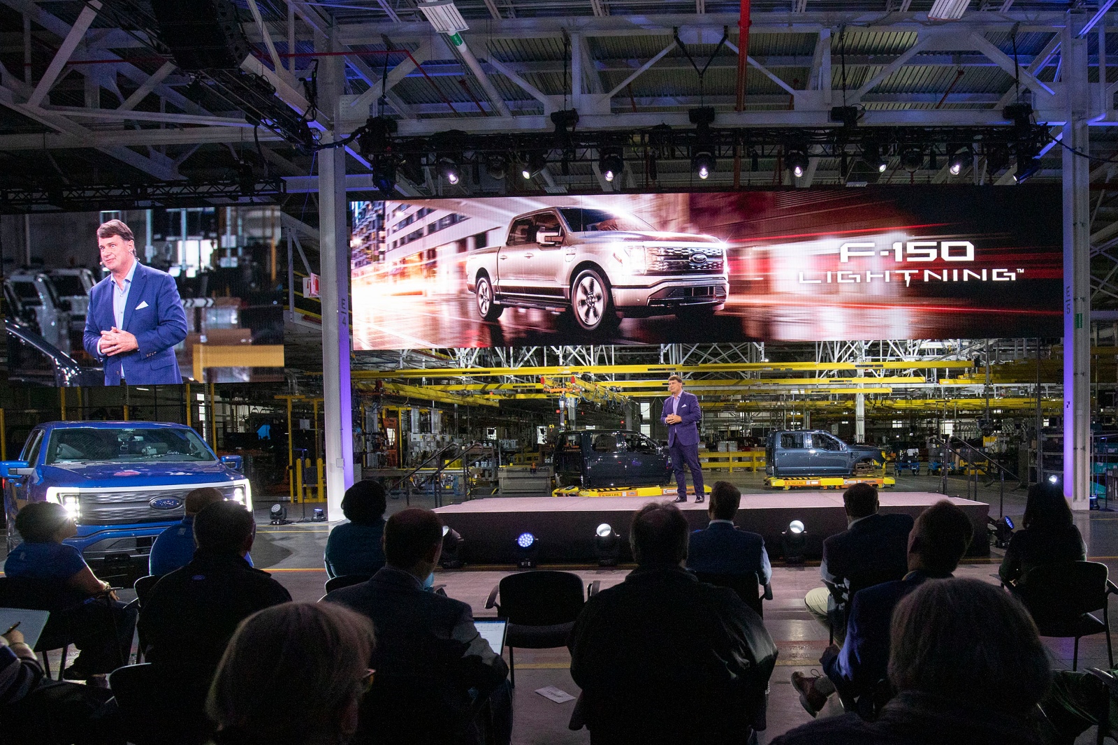 A man in a blue suit stands onstage, next to a Ford pick-up truck, as an audience watches.