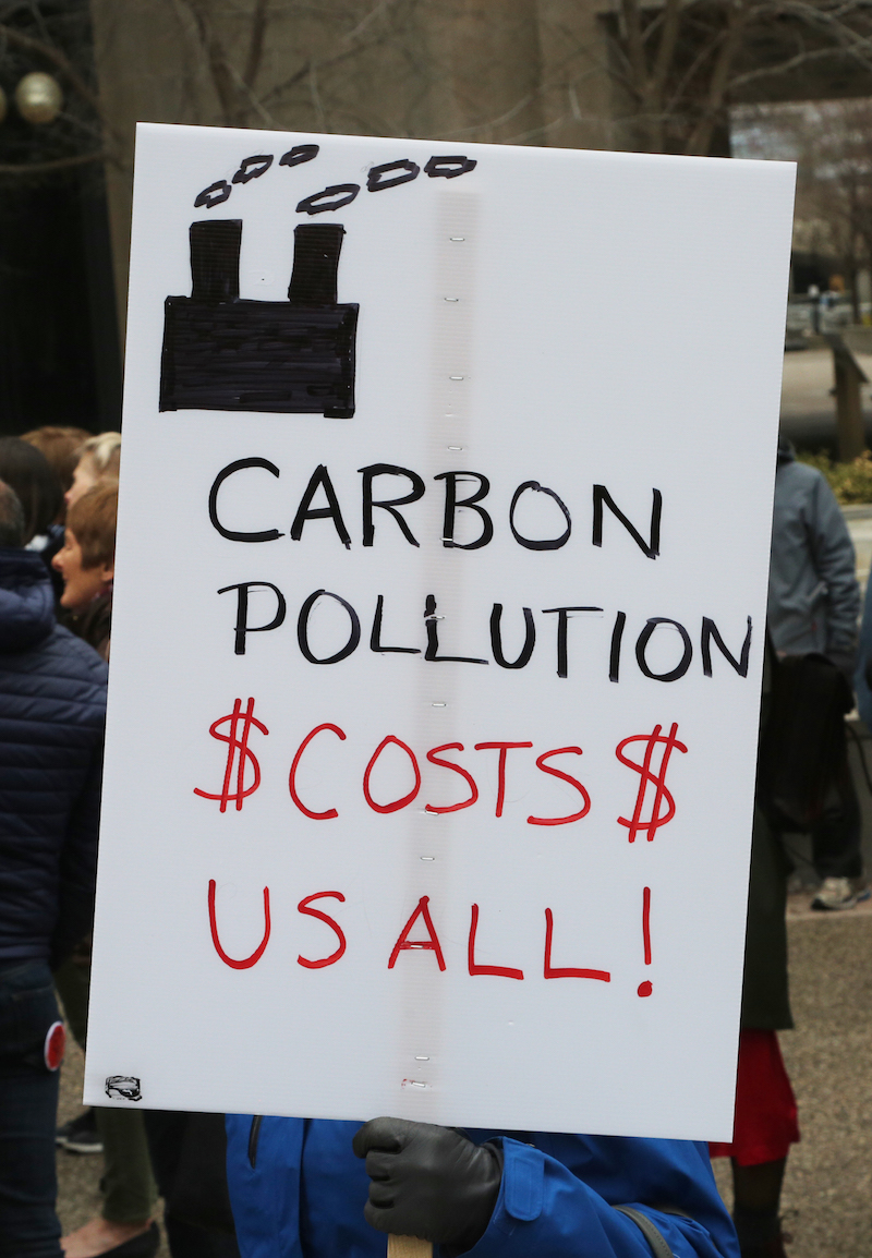 a protester holds a sign that says "carbon pollution $costs$ us all"