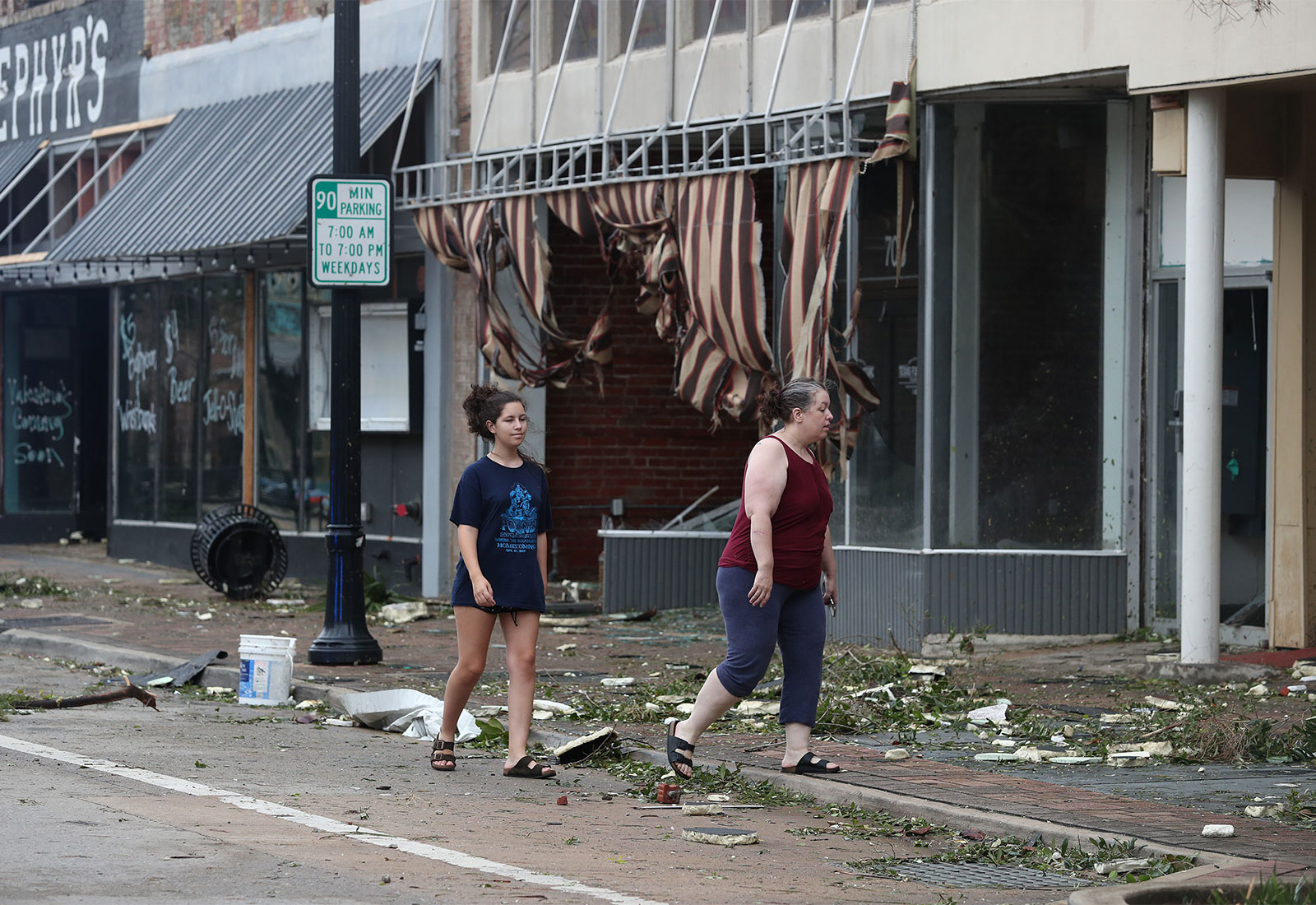 Two women walking on a street in a downtown area of Lake Charles, Louisiana; trash and debris litter the street and shopfronts have torn awnings and shattered windows
