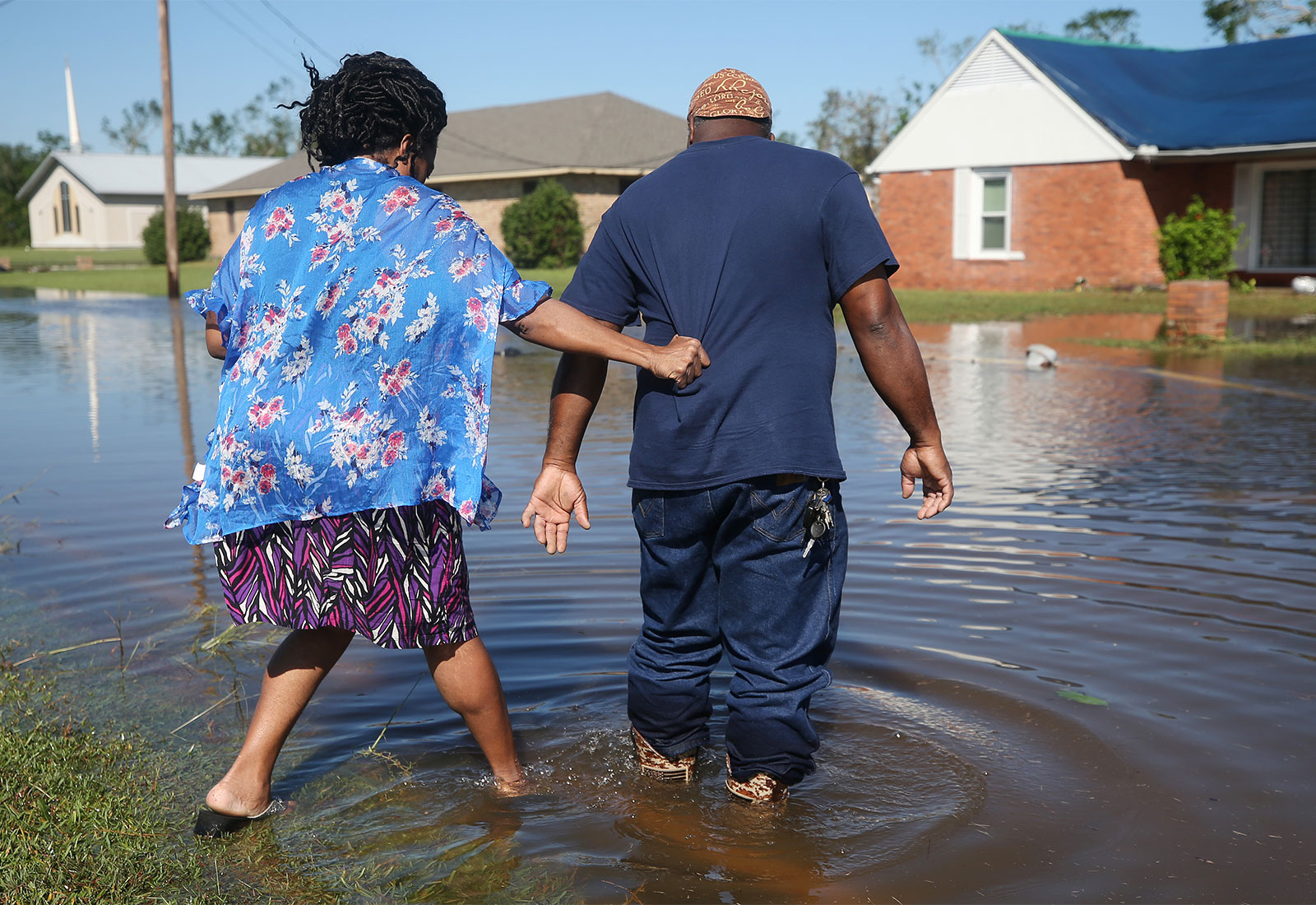 A woman (left) wearing a colorful floral outfit holds onto the back of a man's (right) shirt as they step into a flooded street towards a white and red brick home with a blue tarp on the roof