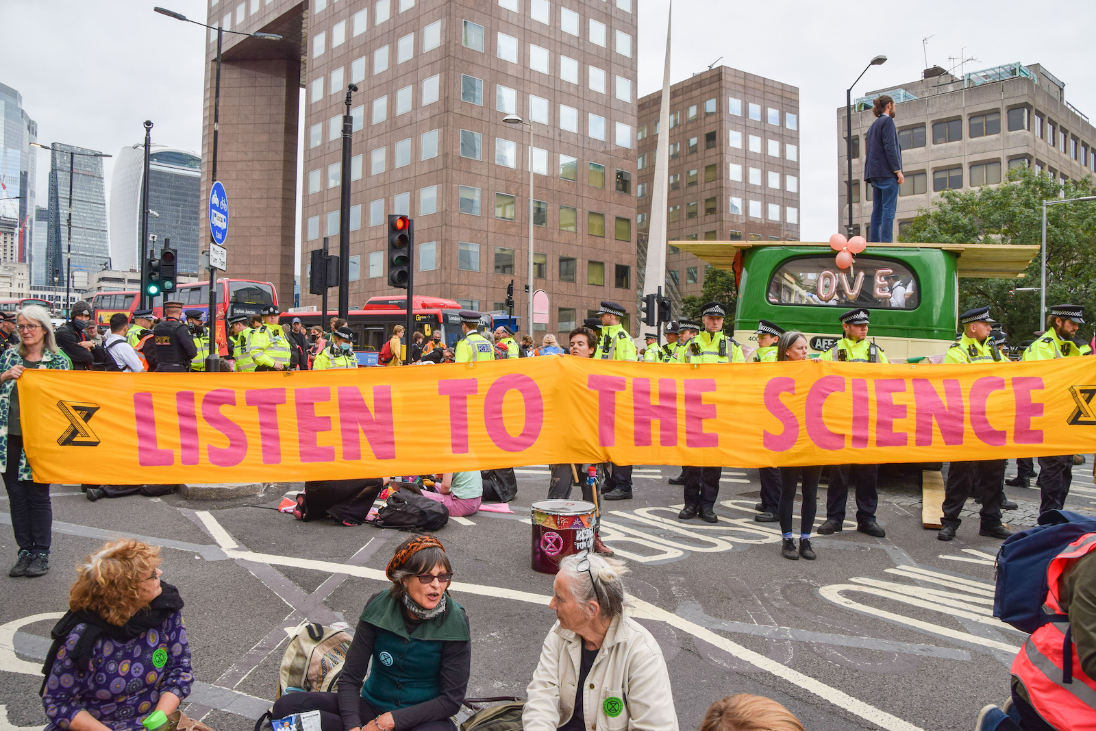 a group of people in neon green police vests stand behind a large yellow and red banner that reads listen to the science