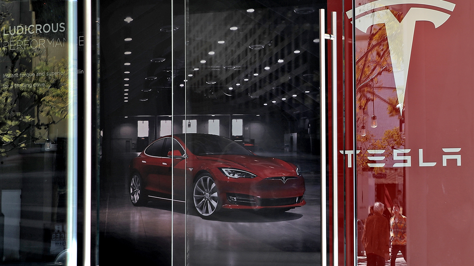 A Tesla showroom seen from the outside; red car and banner with Tesla logo visible through the windows