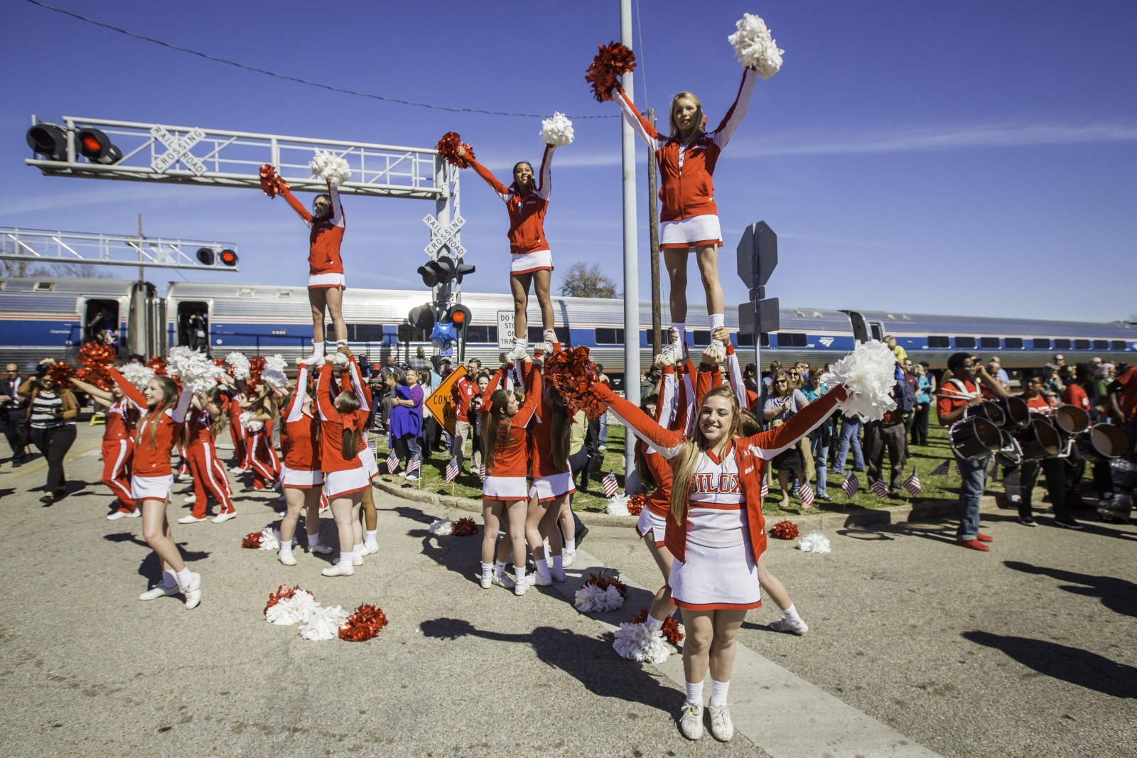 Cheerleaders in red celebrate in front of an Amtrak train at a crowded station event.
