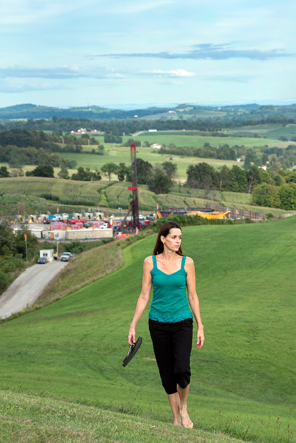 Woman walks up a grassy hill with oil and gas infrastructure in the background