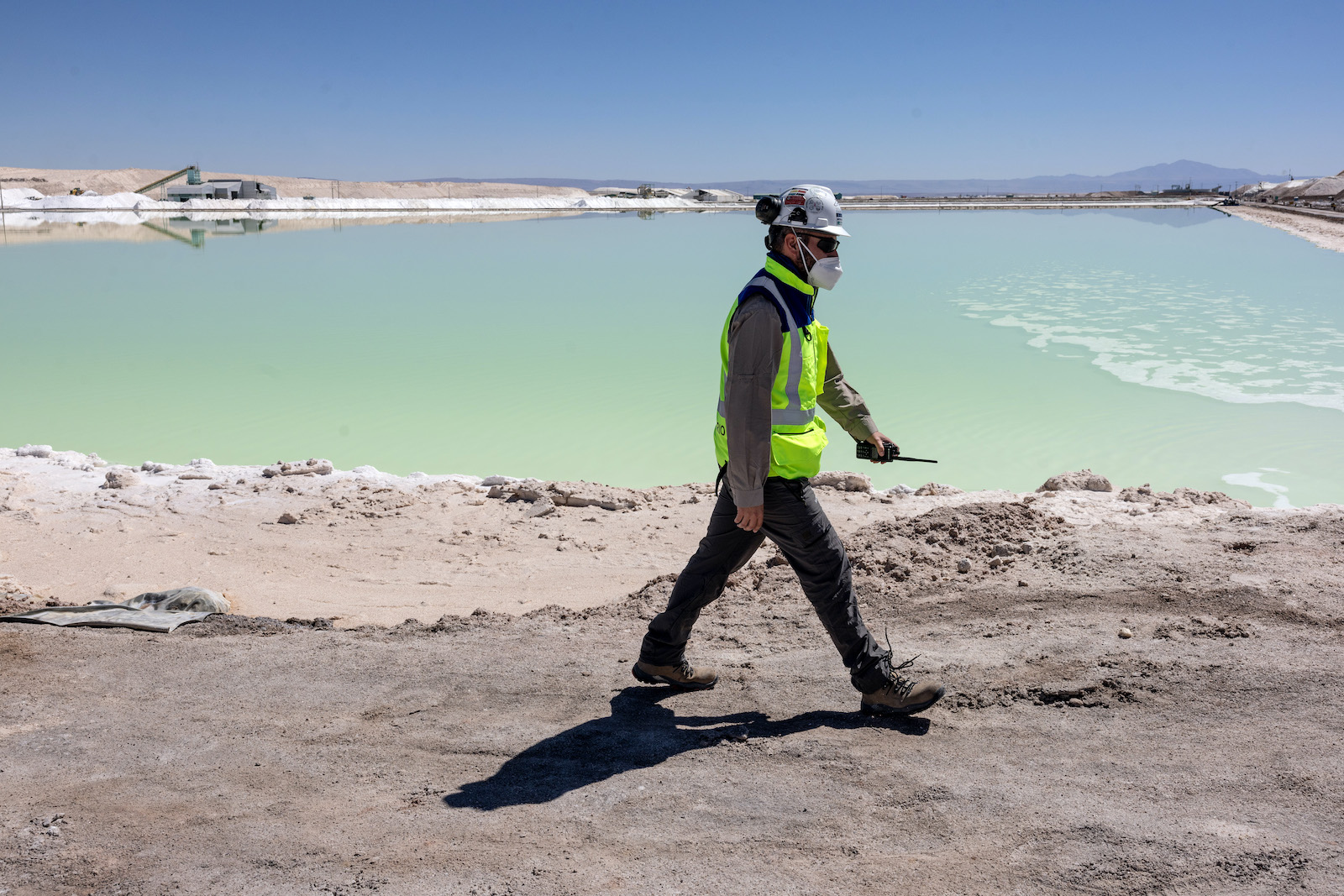 a man in a green vest and hard hat walks past a turqouise lake