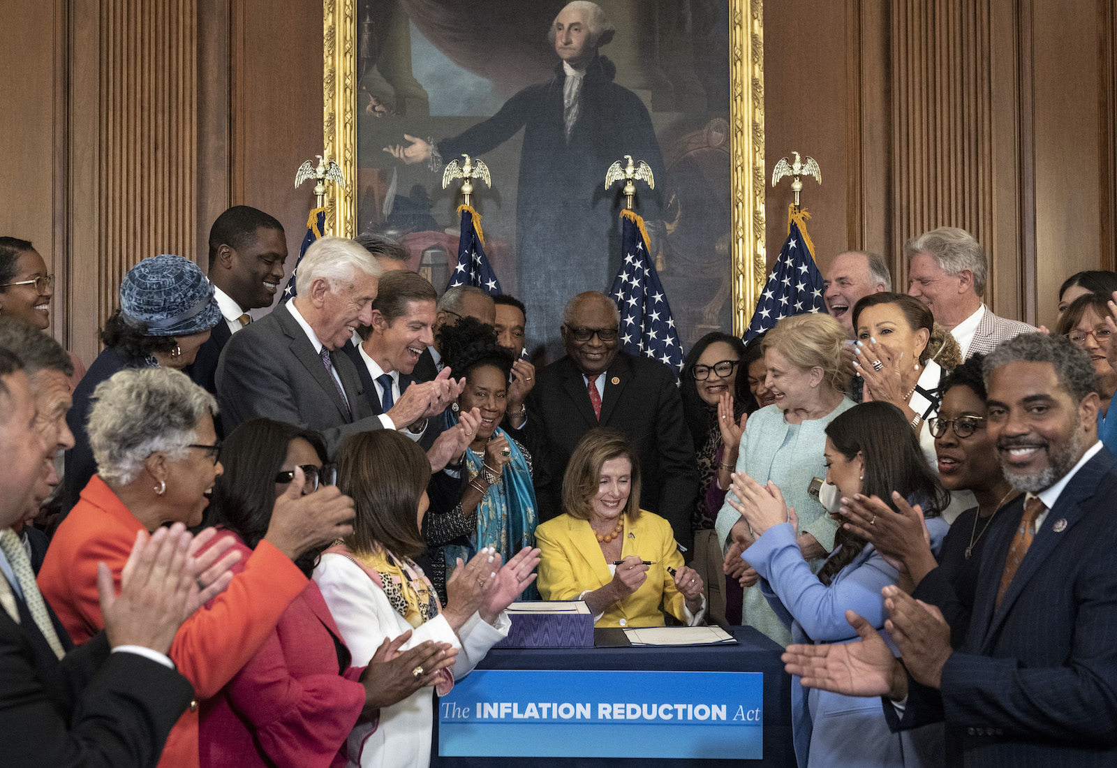 Nancy Pelosi seated in front of a portrait of George Washington, surrounded by lawmakers