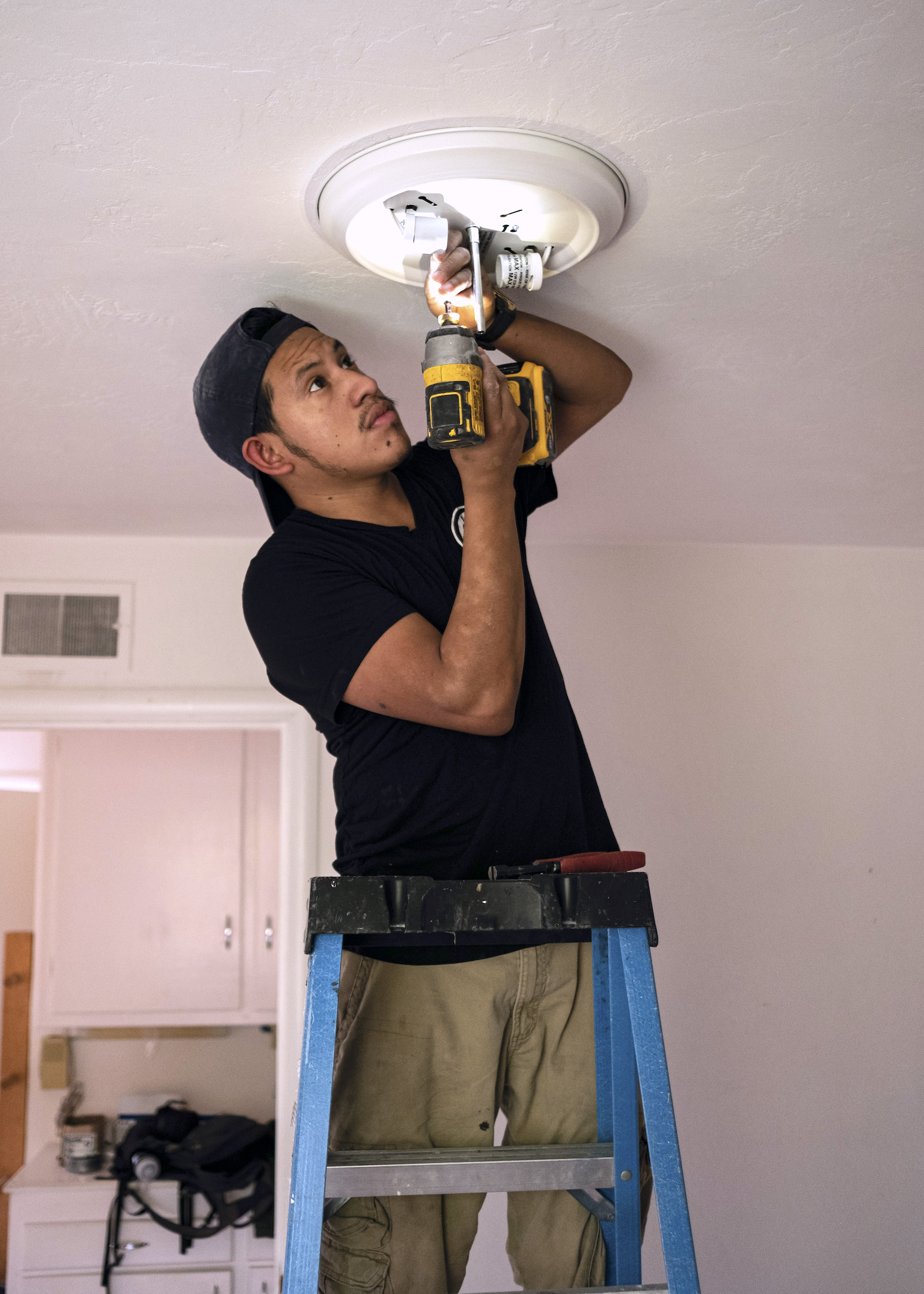 a man in a black t-shirt stands on a blue ladder using tools on a light fixture